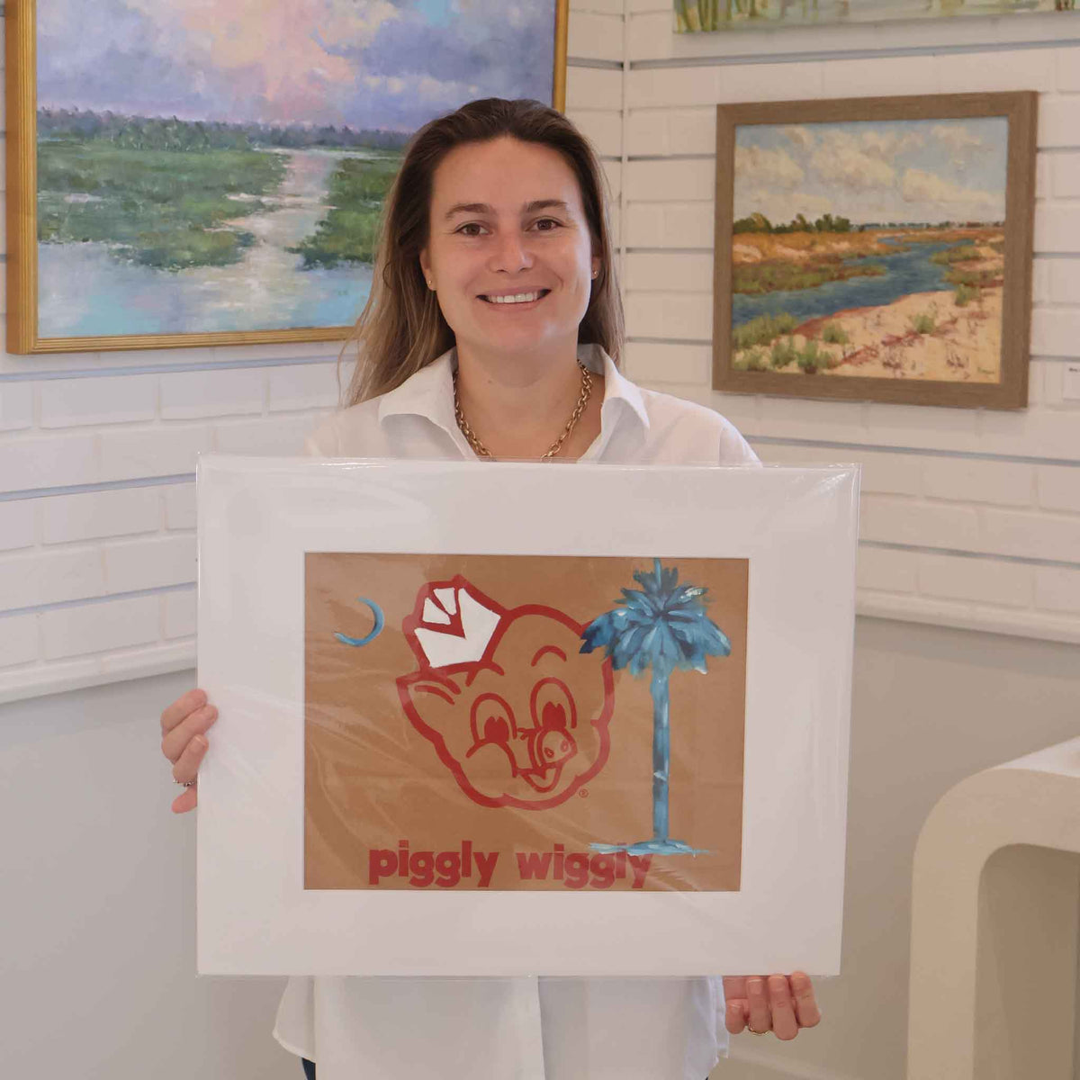 Gray Man Gallery team member holding Carolina on My Mind by local South Carolina artist Jenny Reynolds, an original acrylic painting featuring a vivid blue palmetto tree and crescent moon over the classic red Piggly Wiggly logo on a genuine grocery bag. The matted artwork is photographed inside Gray Man Gallery in Pawleys Island, South Carolina.