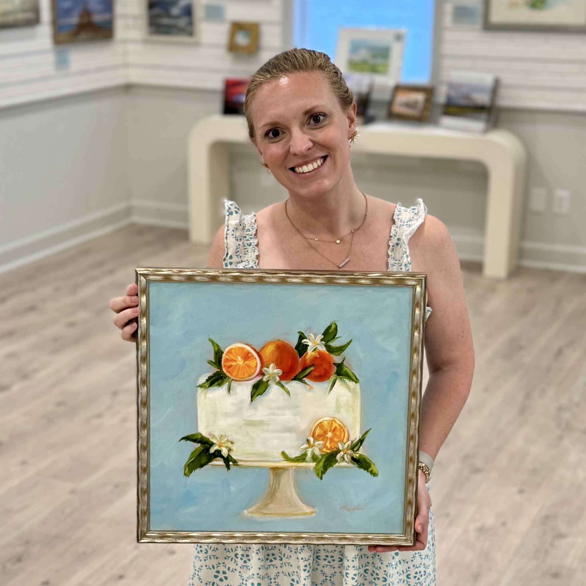 Local artist Jenny Reynolds holding Creamsicle, an original square oil painting featuring a citrus-topped white cake in her Southern Sweets Collection, framed in a signature champagne frame at Gray Man Gallery