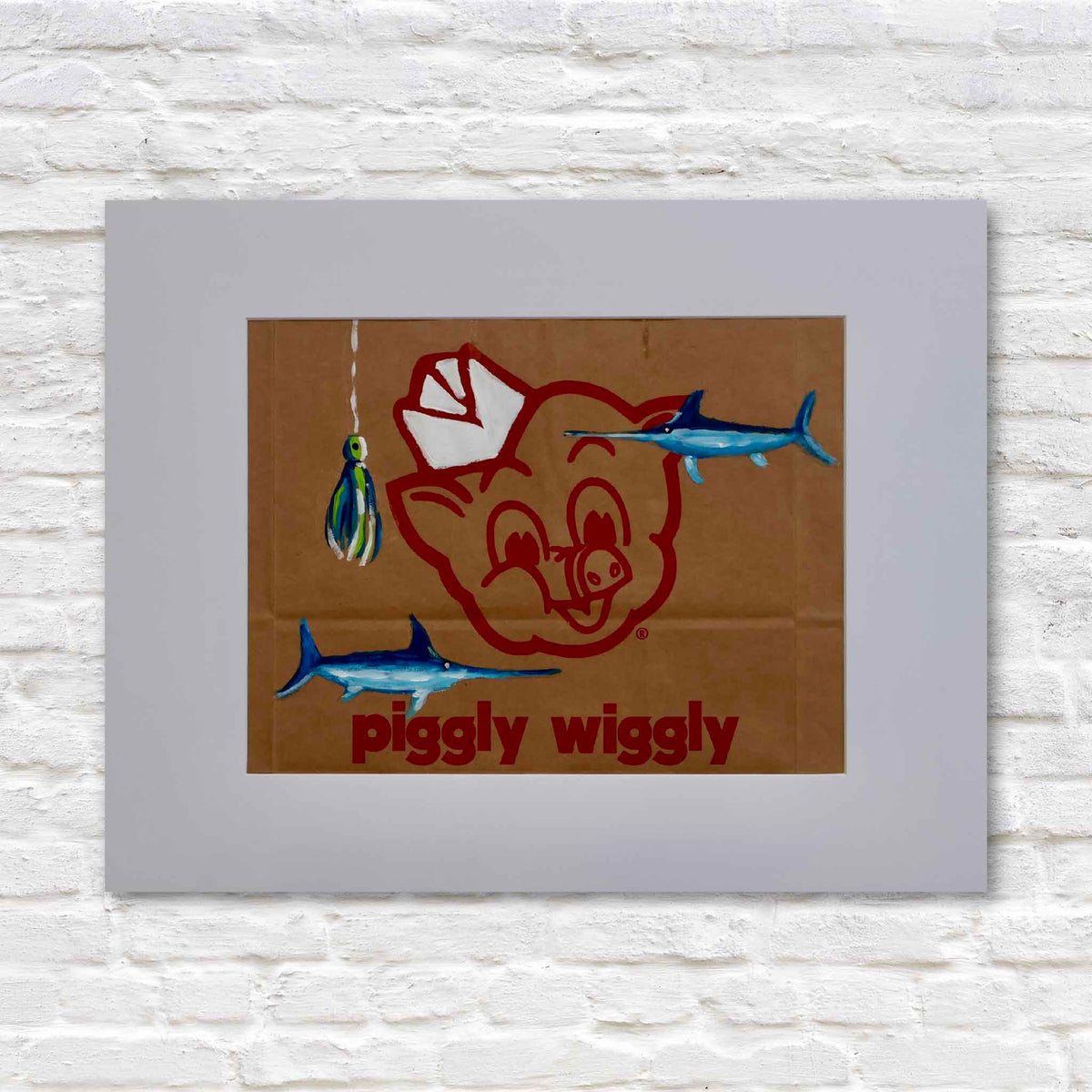 Hook, Line & Piggy, original acrylic painting by local artist Jenny Reynolds featuring two vivid blue saltwater game fish and a colorful hanging fishing lure painted on an authentic brown Piggly Wiggly grocery bag. The recognizable red pig mascot and lettering remain visible beneath the coastal artwork, presented in a wide light gray mat against a white brick background at Gray Man Gallery in Pawleys Island, South Carolina.