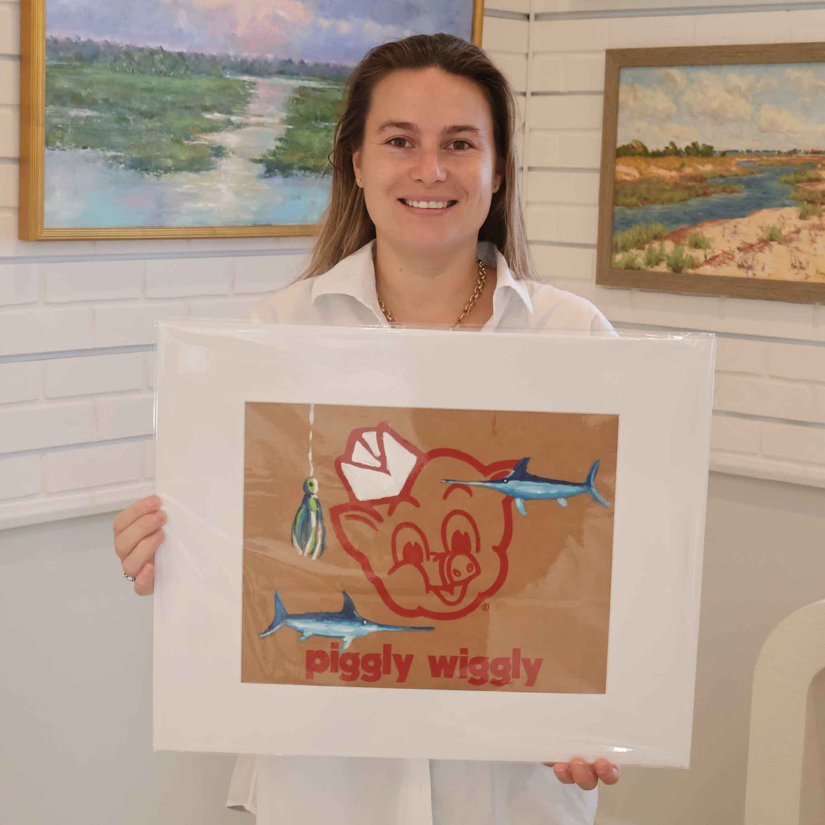 Gray Man Gallery team member holding Hook, Line & Piggy, an original acrylic painting by local artist Jenny Reynolds featuring two blue saltwater fish and a colorful fishing lure over the classic red Piggly Wiggly logo. The one-of-a-kind artwork is painted on a genuine grocery bag and displayed in a wide mat inside Gray Man Gallery in Pawleys Island, South Carolina.