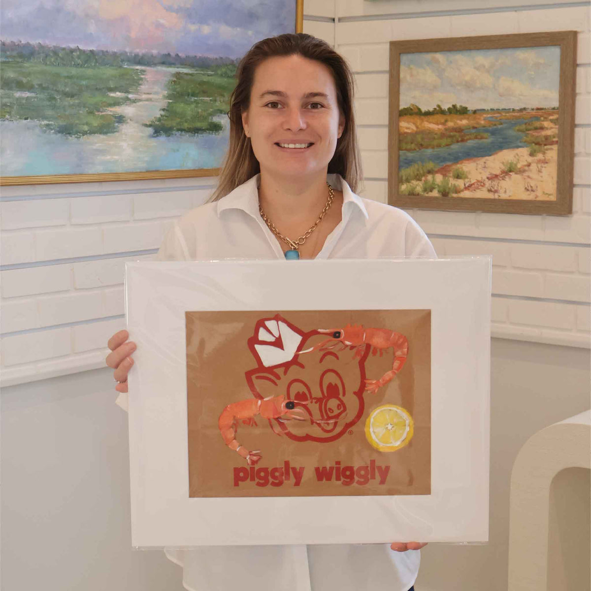 Gray Man Gallery team member holding Peel & Eat by local South Carolina artist Jenny Reynolds, a one-of-a-kind original acrylic painting of two shrimp and a lemon on an authentic Piggly Wiggly grocery bag. Shown with its wide white mat inside Gray Man Gallery in Pawleys Island.
