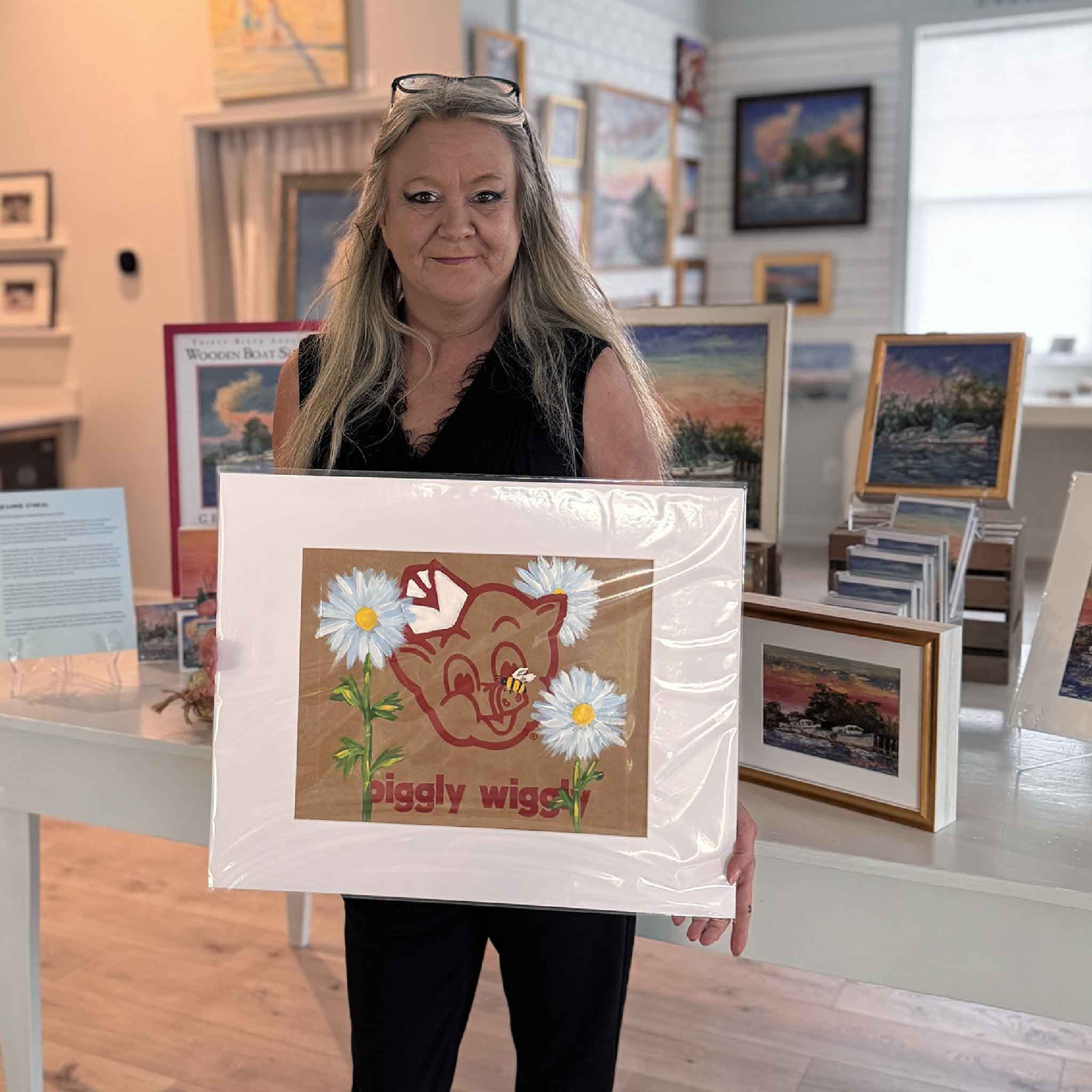 Charlotte holding “Piggly Wiggly: Daisies & Bee,” an original Jenny Reynolds painting on a Piggly Wiggly grocery bag featuring white daisies and a bee.