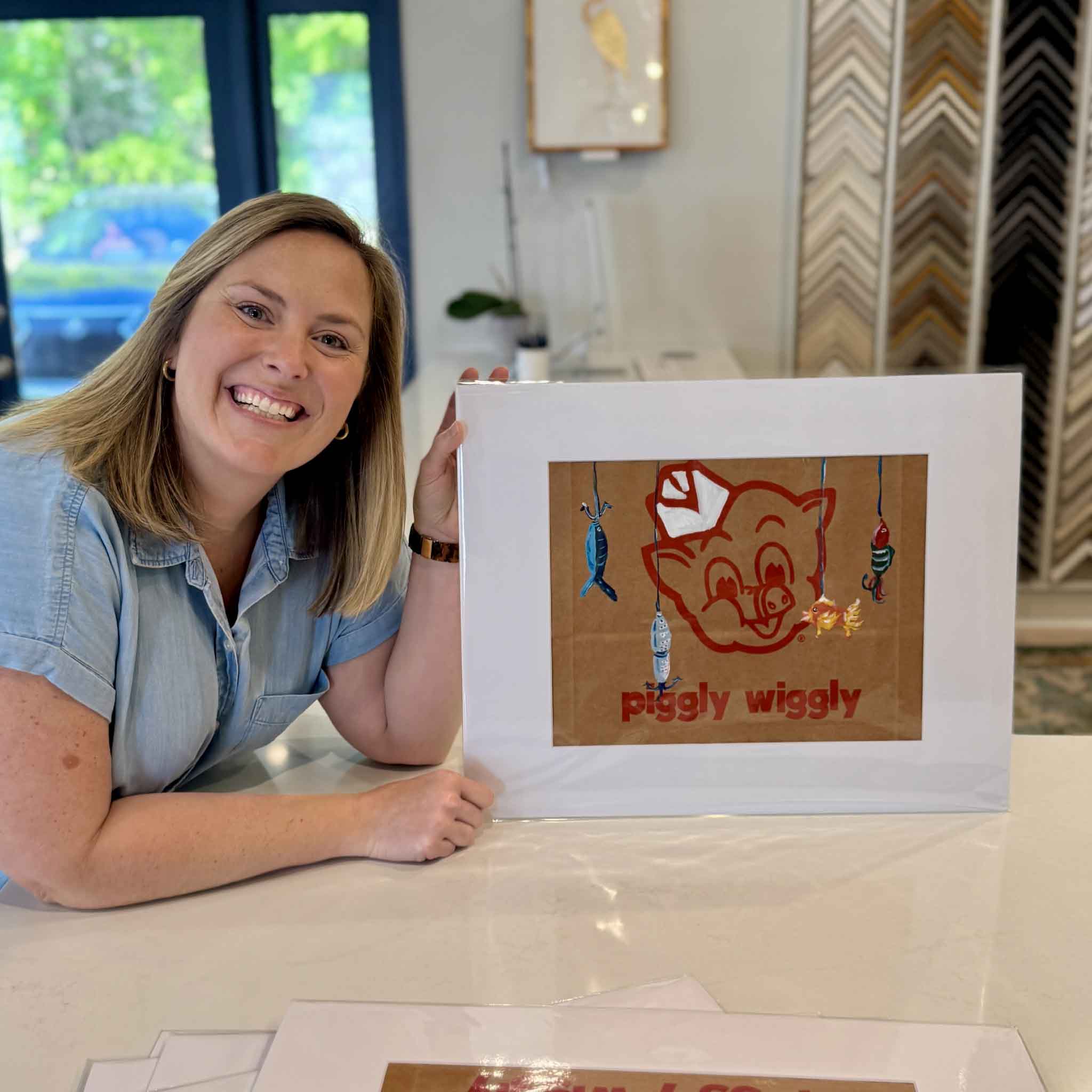 Team member holding Piggly Wiggly: Fishin’ Lures by Jenny Reynolds, an original mixed media painting on a Piggly Wiggly grocery bag matted to fit a standard 16x20 frame at Gray Man Gallery in Pawleys Island