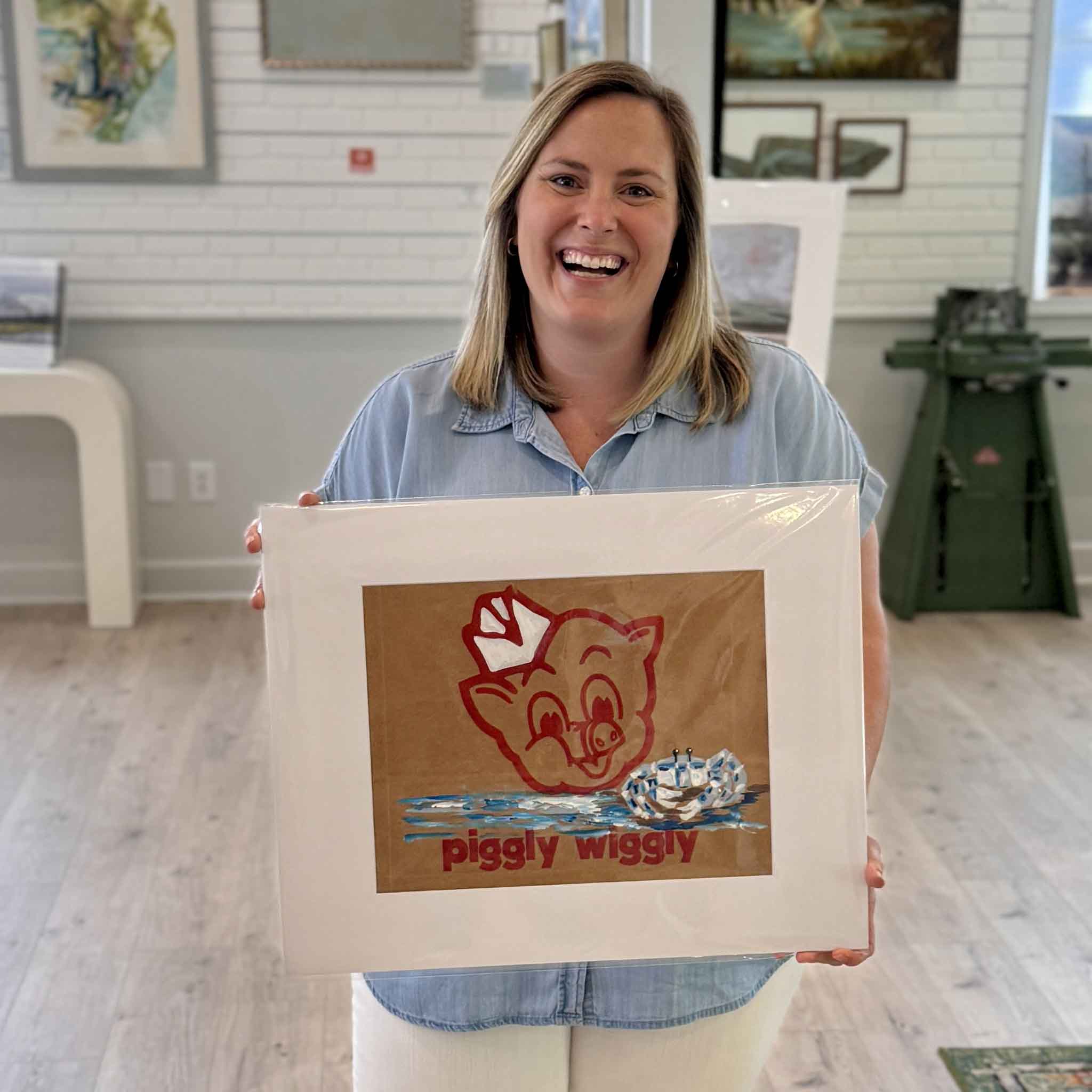 Bordy holding an original Piggly Wiggly Ghost Crab painting by local artist Jenny Reynolds inside Gray Man Gallery, showing scale of the hand-painted grocery bag artwork with coastal crab detail and iconic pig logo