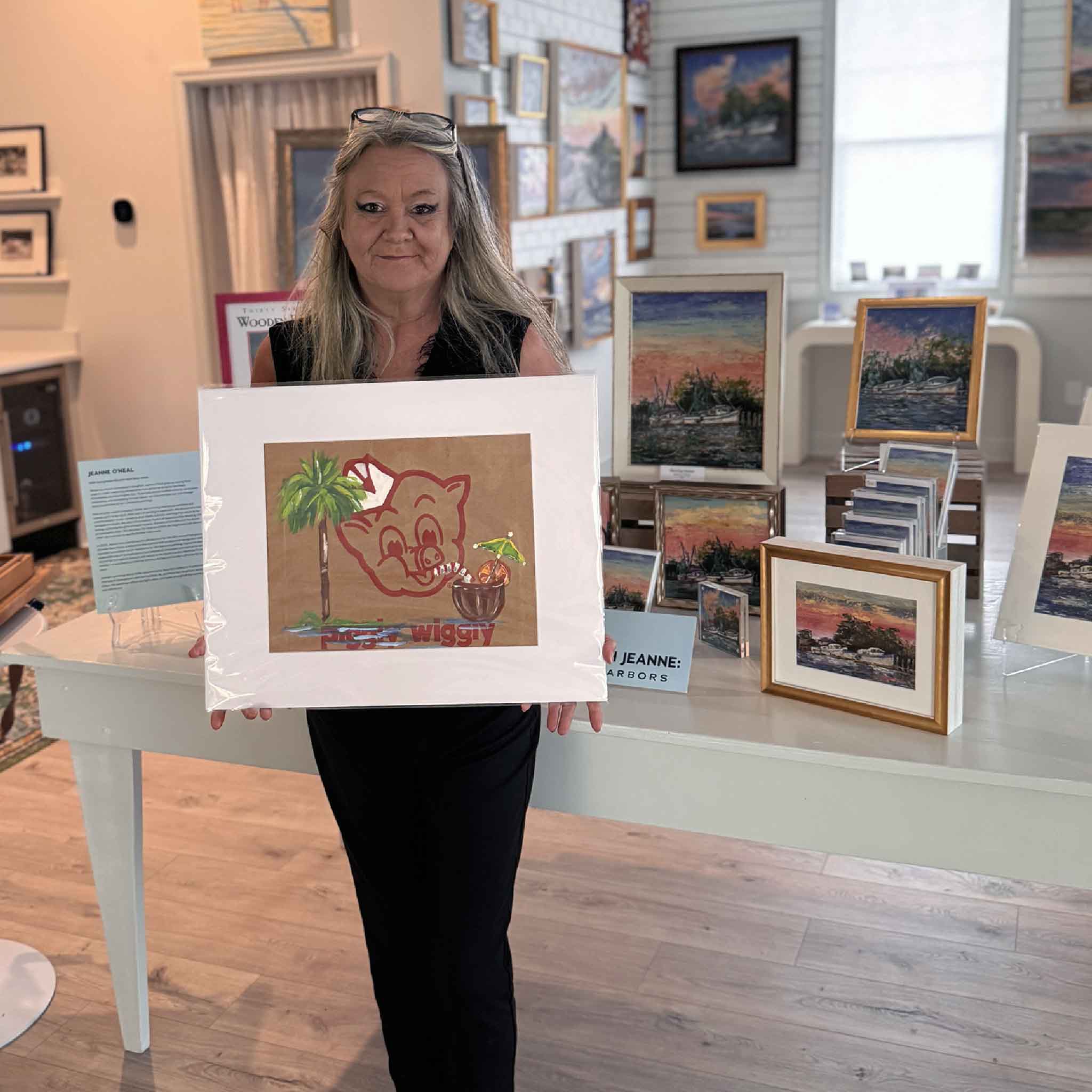 Charlotte holding “Piggly Wiggly: Piña Colada,” an original Jenny Reynolds painting on a Piggly Wiggly grocery bag featuring a palm tree, pig logo, and tropical drink.