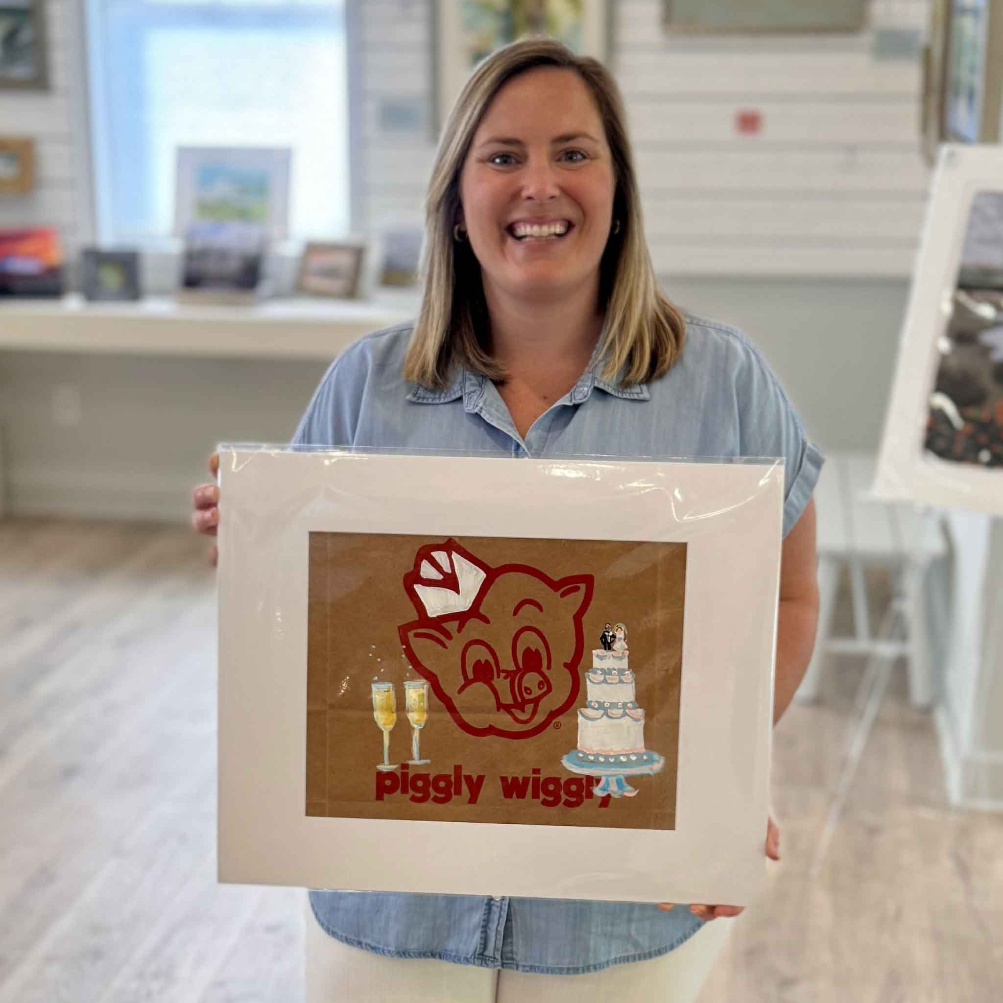 Bordy holding an original Piggly Wiggly Wedding Bells painting by local artist Jenny Reynolds inside Gray Man Gallery, showing scale of the hand-painted grocery bag artwork with champagne glasses and wedding cake detail