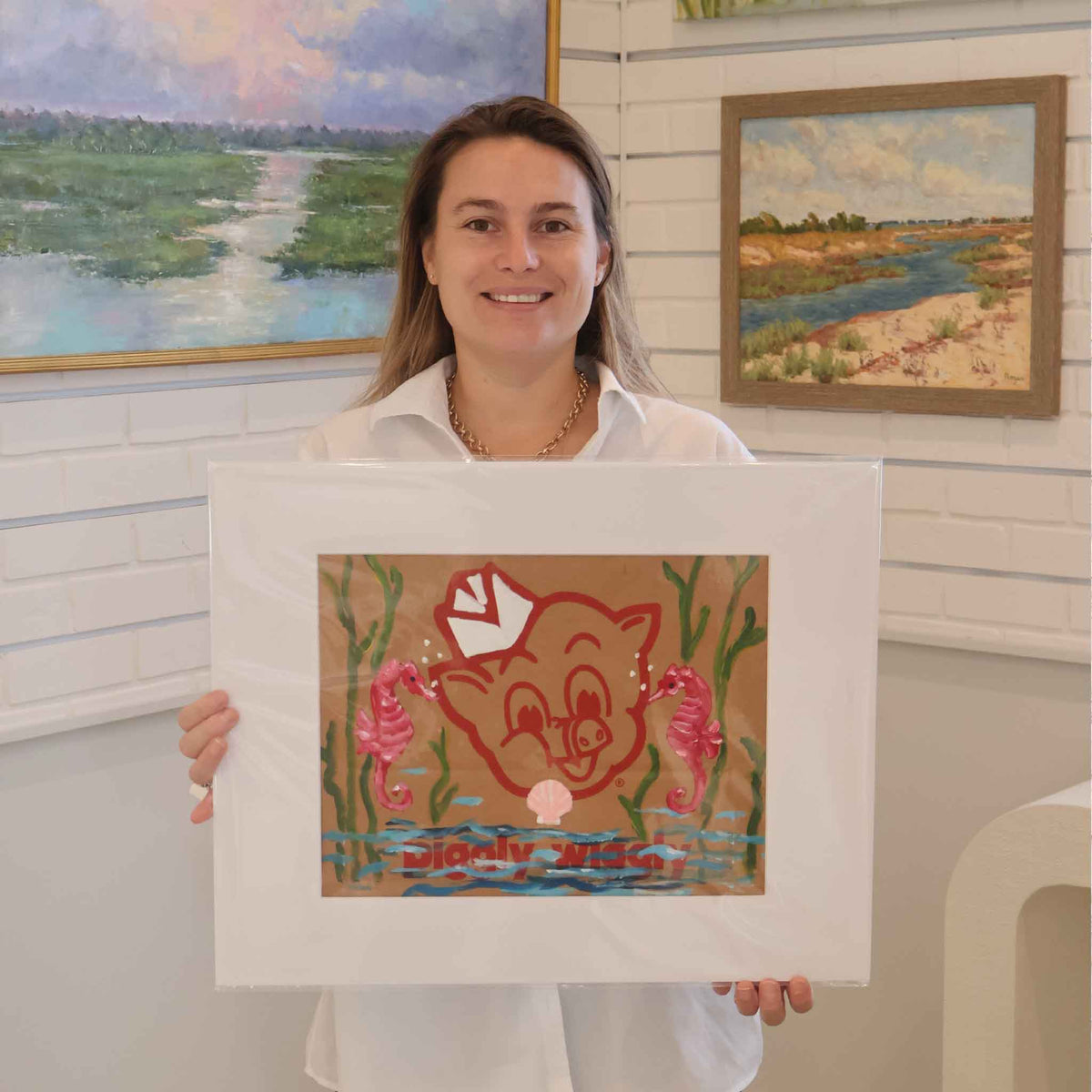 Gray Man Gallery team member holding Seahorsin’ Around by local artist Jenny Reynolds, a one-of-a-kind original acrylic painting of pink seahorses, underwater plants, and blue waves over the classic red Piggly Wiggly logo on an authentic grocery bag. The matted coastal artwork is photographed inside Gray Man Gallery in Pawleys Island, South Carolina.