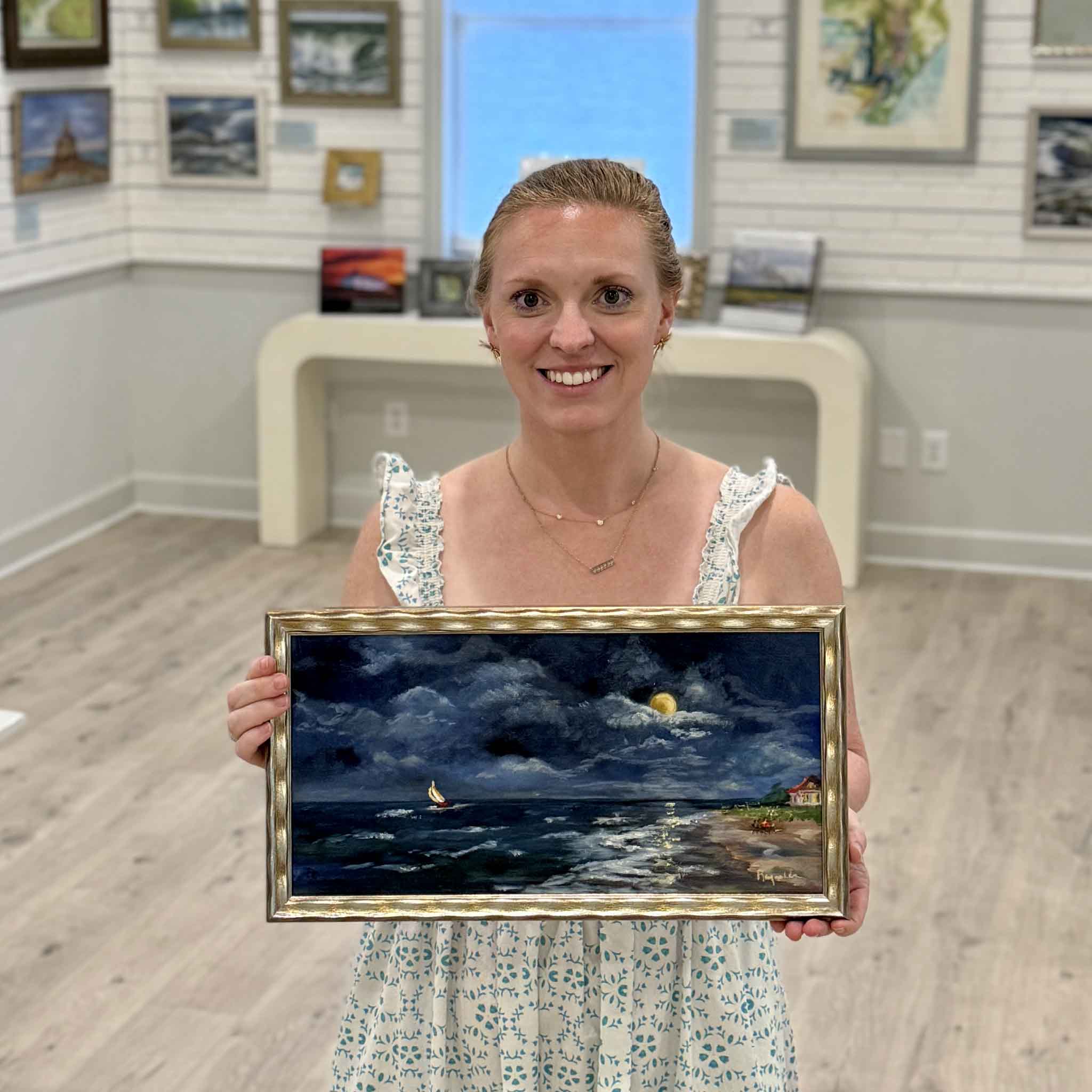 Local artist Jenny Reynolds holding Summer Nights, an original moonlit coastal oil painting with crashing waves, deep blue skies, and glowing shoreline light, framed in her signature gold frame at Gray Man Gallery in Pawleys Island