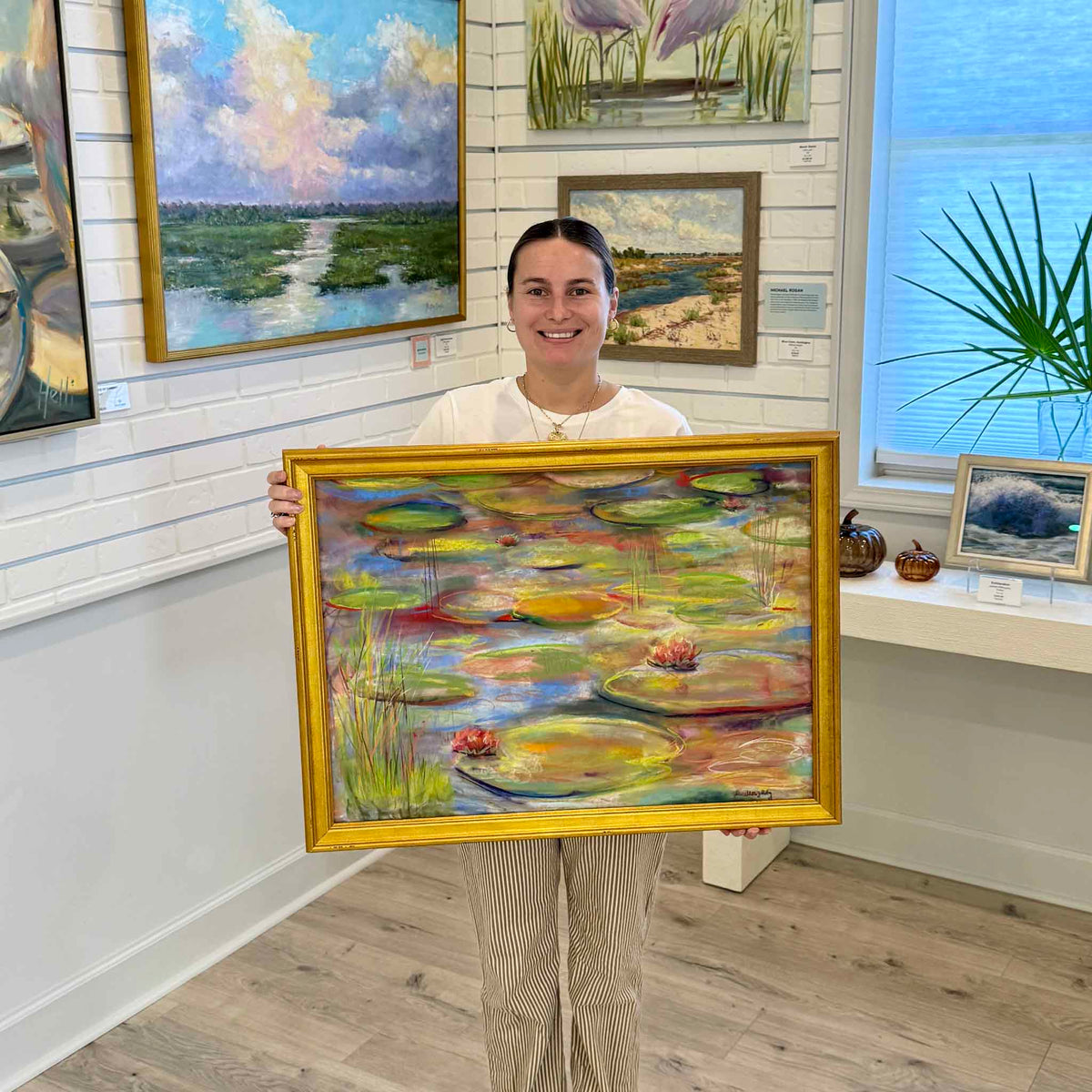 Framed original pastel painting Brookgreen Beauty by local artist Joanne Willoughby, held by a Gray Man Gallery team member inside the Pawleys Island, South Carolina art gallery. The colorful Brookgreen Gardens-inspired lily pond painting features floating green lily pads, pink water lilies, vibrant reflections, and a traditional gold frame, showing the scale and presentation of the original artwork.