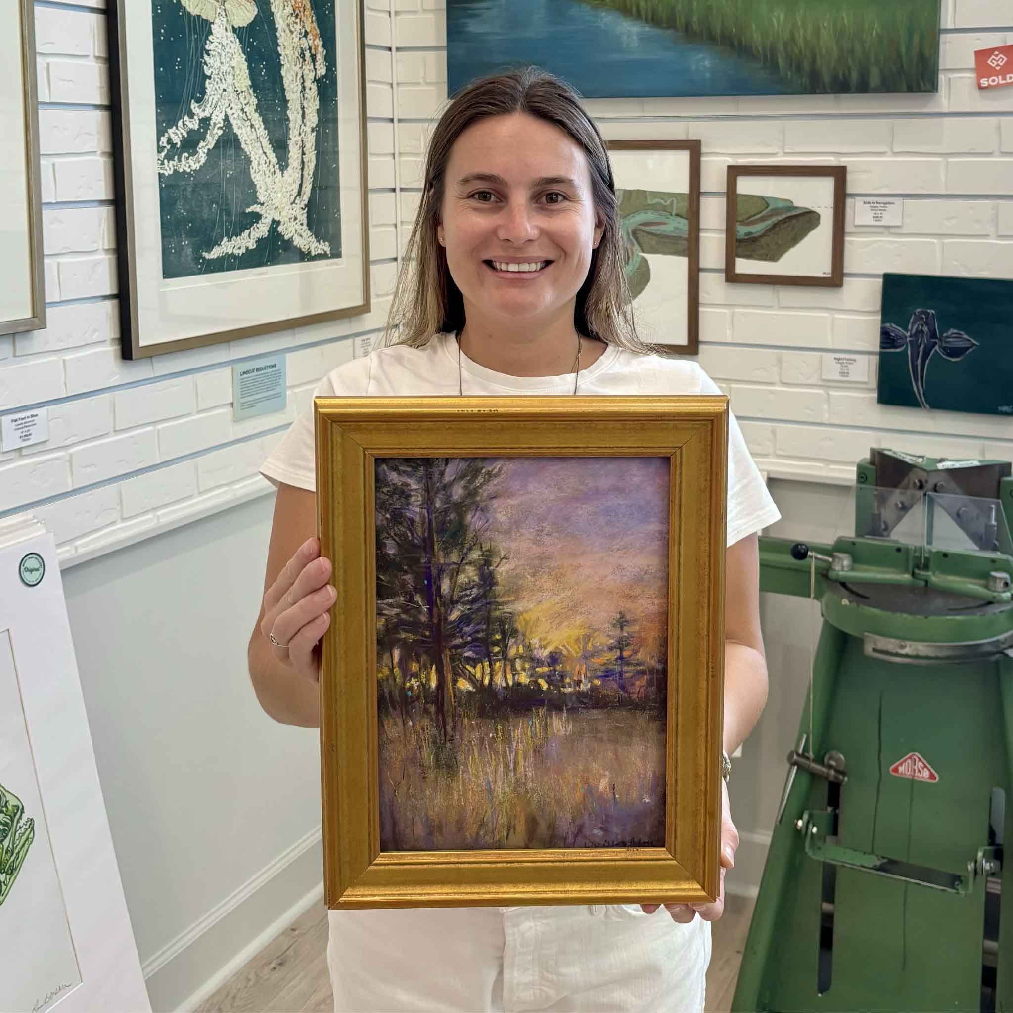 Kyra holds Forest of Dreams, an original pastel painting by artist Joanne Willoughby, at Gray Man Gallery in Pawleys Island, South Carolina, featuring an atmospheric woodland landscape with glowing sunset light, trees and colorful reflections on the water.