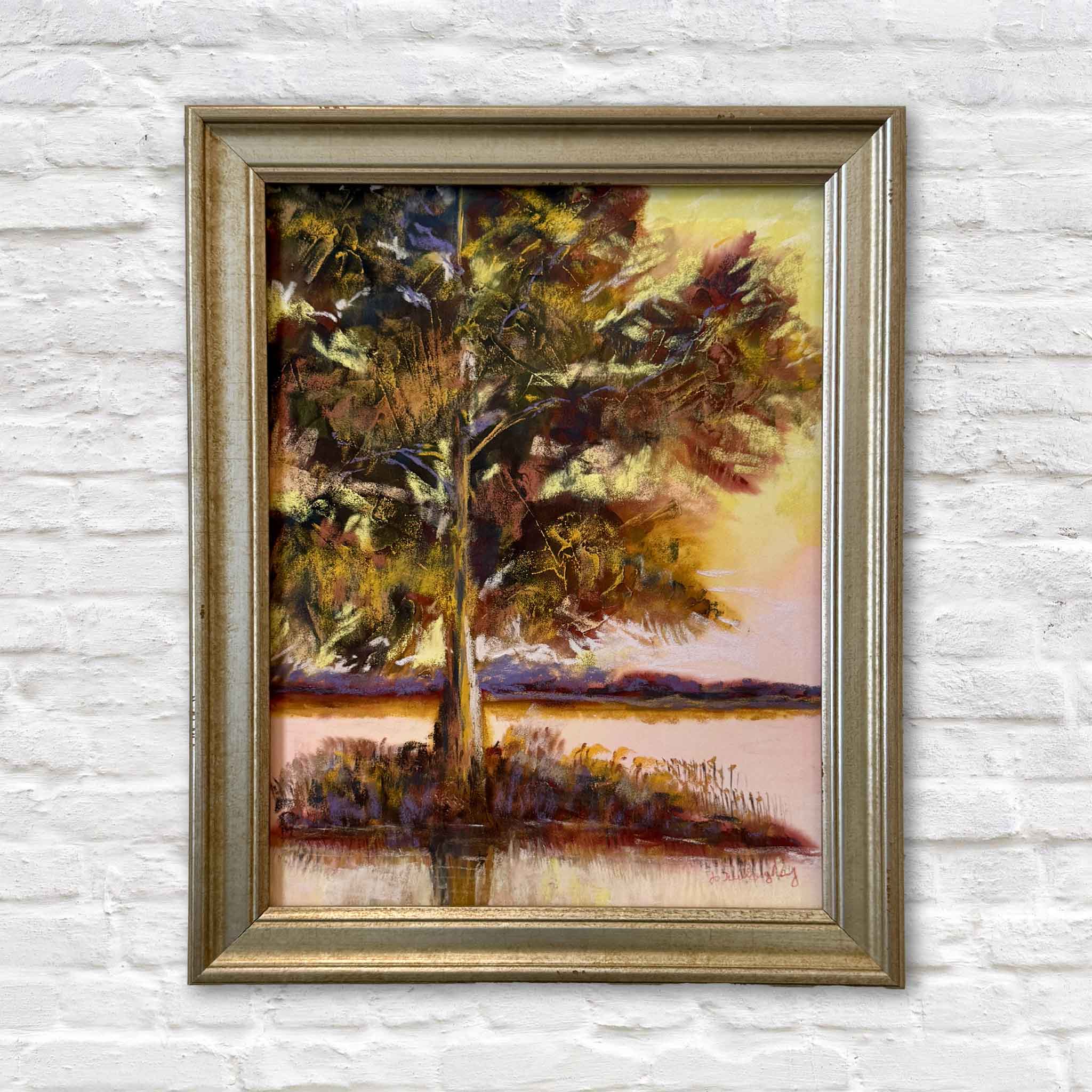 Waccamaw Gold II, an original pastel painting by South Carolina artist Joanne Willoughby, featuring a dramatic tree along the Waccamaw beneath a glowing gold, amber, blush and violet sky. Professionally framed in a warm antiqued metallic frame and photographed at Gray Man Gallery in Pawleys Island, South Carolina, this richly colored original Lowcountry landscape captures the still water, wooded shoreline and atmospheric evening light of the Waccamaw.