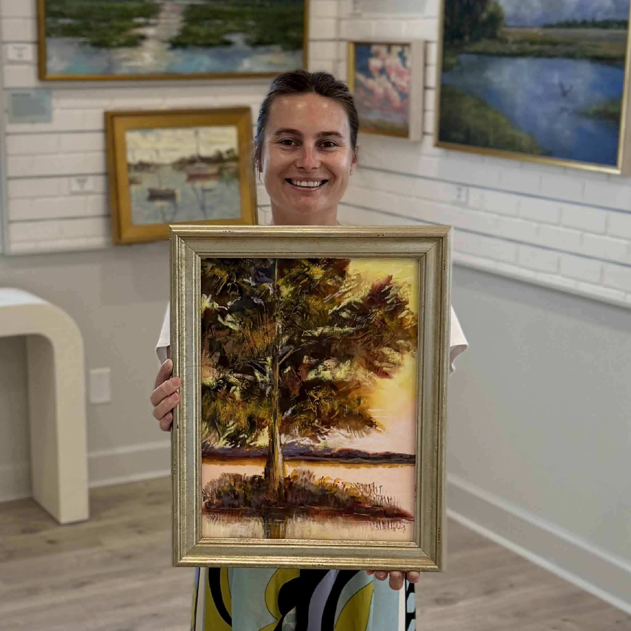 Woman holding Waccamaw Gold II, an original framed pastel painting by South Carolina artist Joanne Willoughby, inside Gray Man Gallery in Pawleys Island for size and scale reference. The vertical Lowcountry landscape features an expressive tree beside the Waccamaw rendered in deep burgundy, olive, gold and violet against luminous evening light, shown professionally framed and surrounded by original coastal artwork in the Pawleys Island gallery.