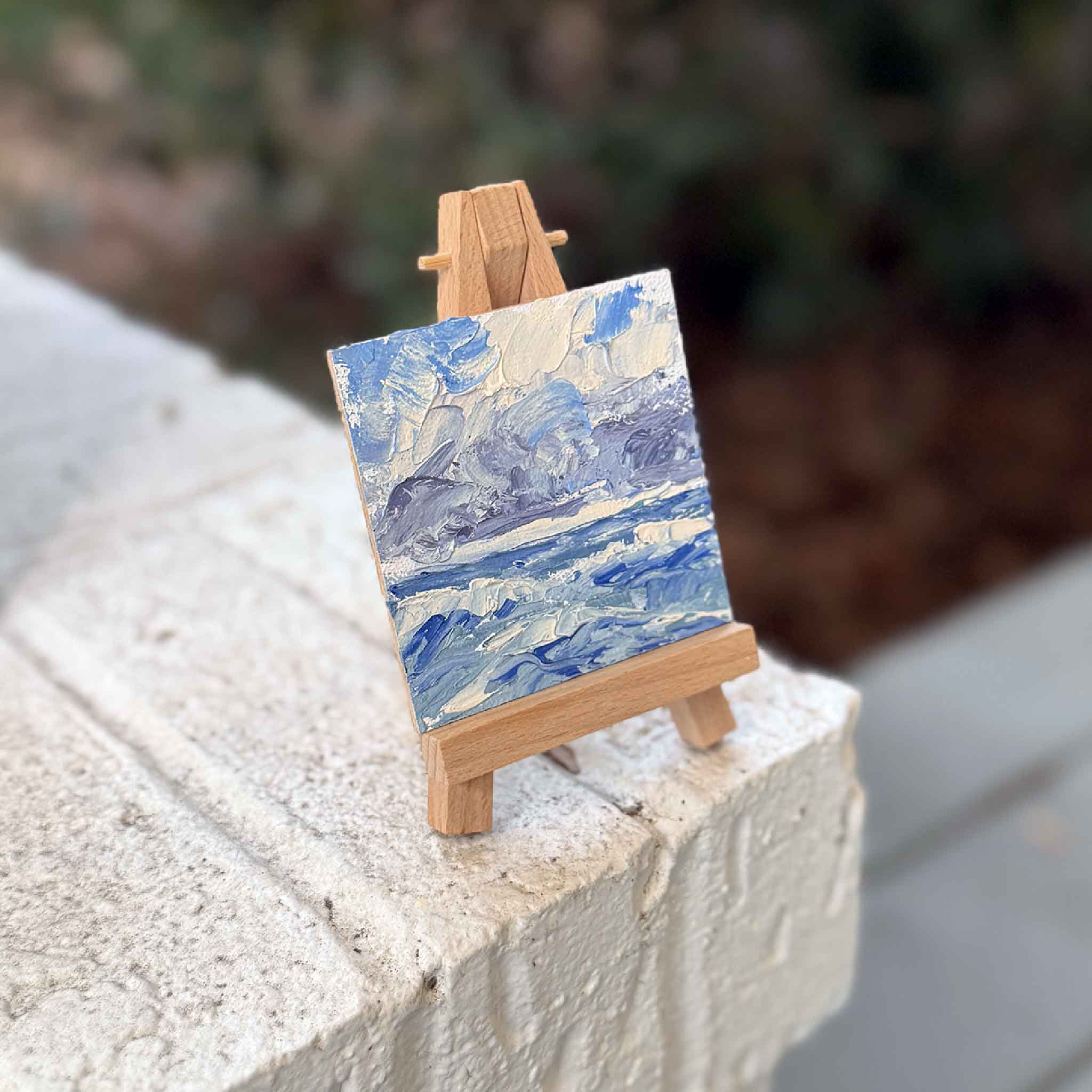 Small coastal oil painting with blue surf and breezy sky by Joanne Willoughby on a tiny wooden easel, photographed outdoors at Gray Man Gallery.