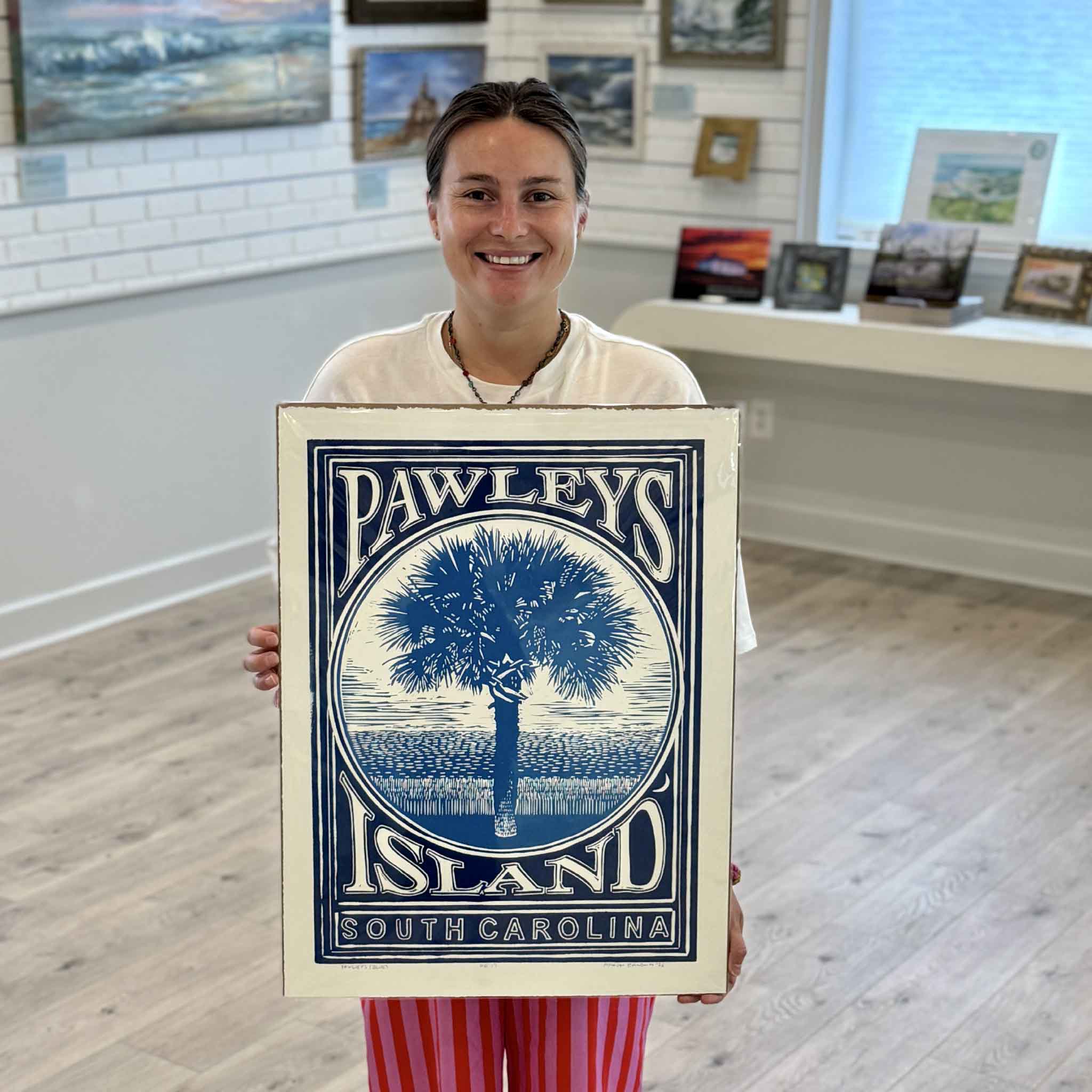 Kyra of Gray Man Gallery holding the blue edition of Aaron Baldwin's Pawleys Island linocut, featuring a palmetto tree and Lowcountry marsh landscape inspired by Pawleys Island, South Carolina.