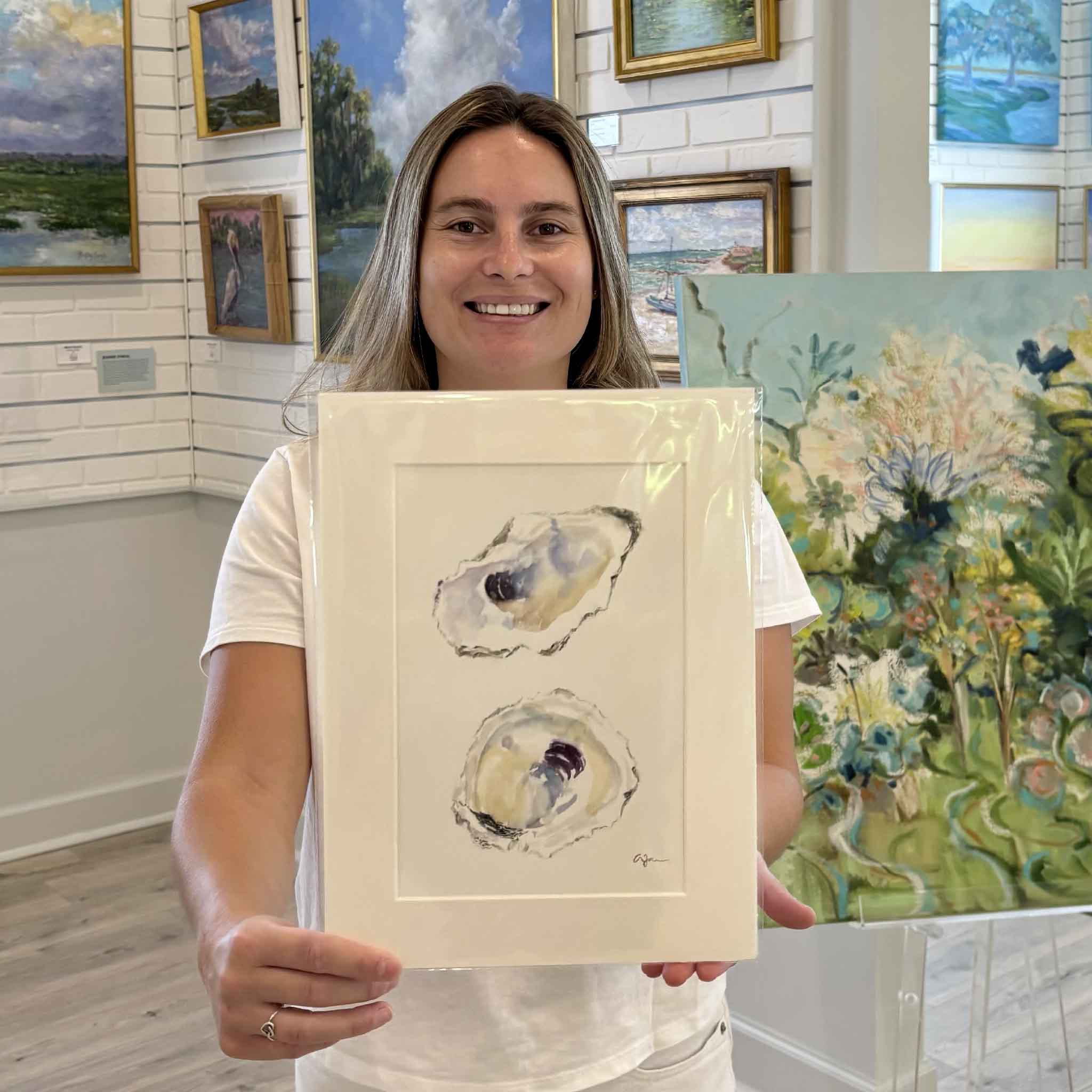 Kyra holding "Serendipity," an original oyster watercolor by Allison Farr from her Lowcountry Jewels series inside Gray Man Gallery in Pawleys Island, South Carolina. The professionally matted original artwork features two delicately painted oyster shells and is ready to fit any standard 9x12 frame.