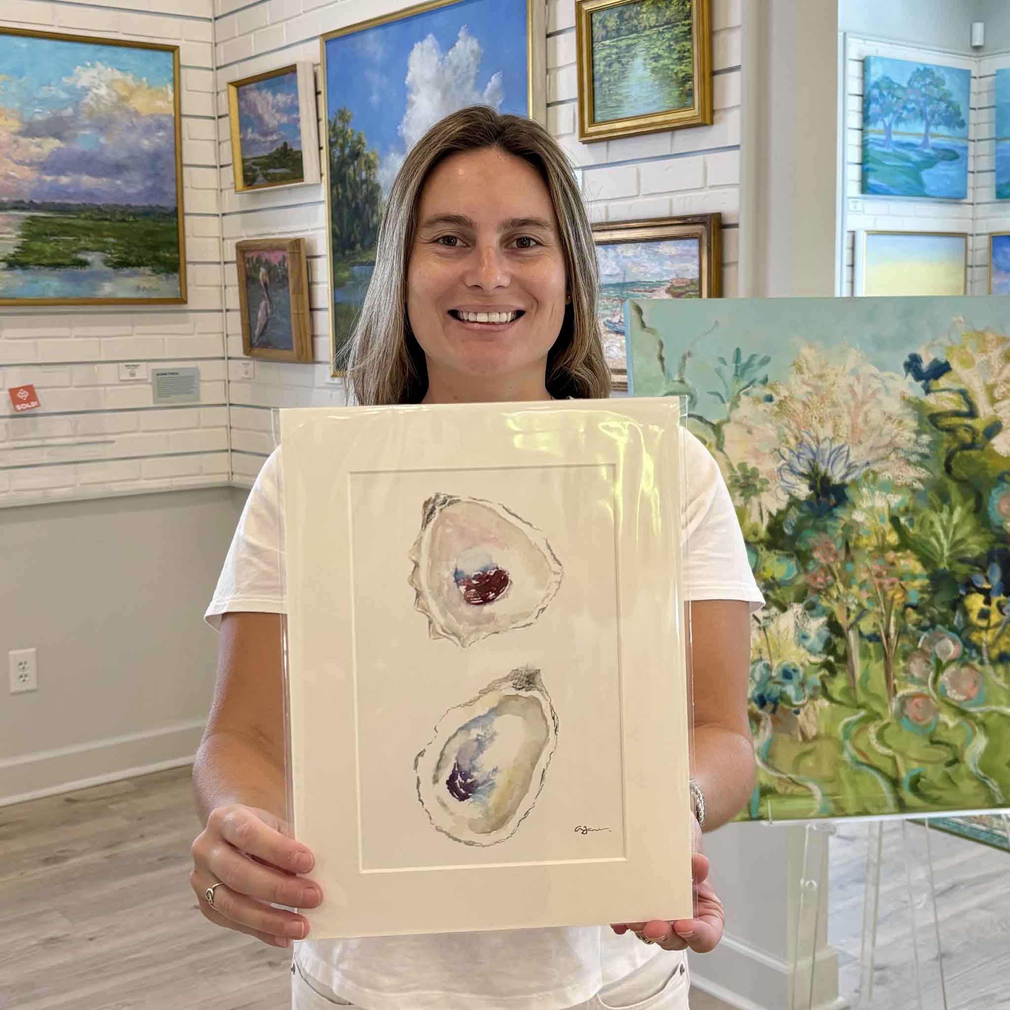 Kyra holding "Stillness," an original oyster watercolor by Allison Farr from her Lowcountry Jewels series inside Gray Man Gallery in Pawleys Island, South Carolina. The professionally matted original artwork features two delicately painted oyster shells and is designed to fit any standard 9x12 frame without custom framing.