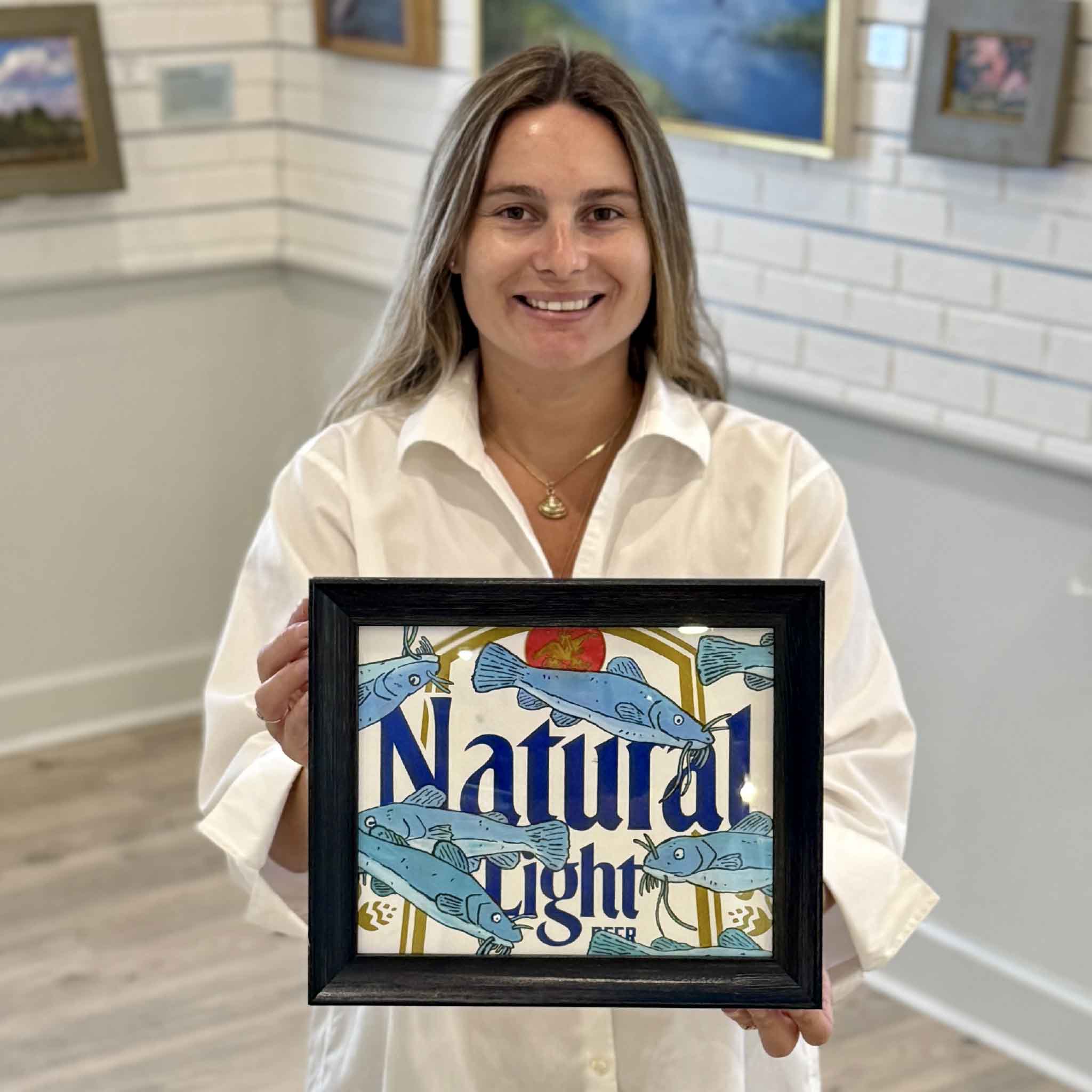 Kyra holds Catties 'n Nattys, an original mixed media artwork by Charleston artist Nick Kawczynski featuring hand-painted catfish on a reclaimed Natural Light beer box. Professionally framed in navy blue and available at Gray Man Gallery in Pawleys Island, South Carolina.