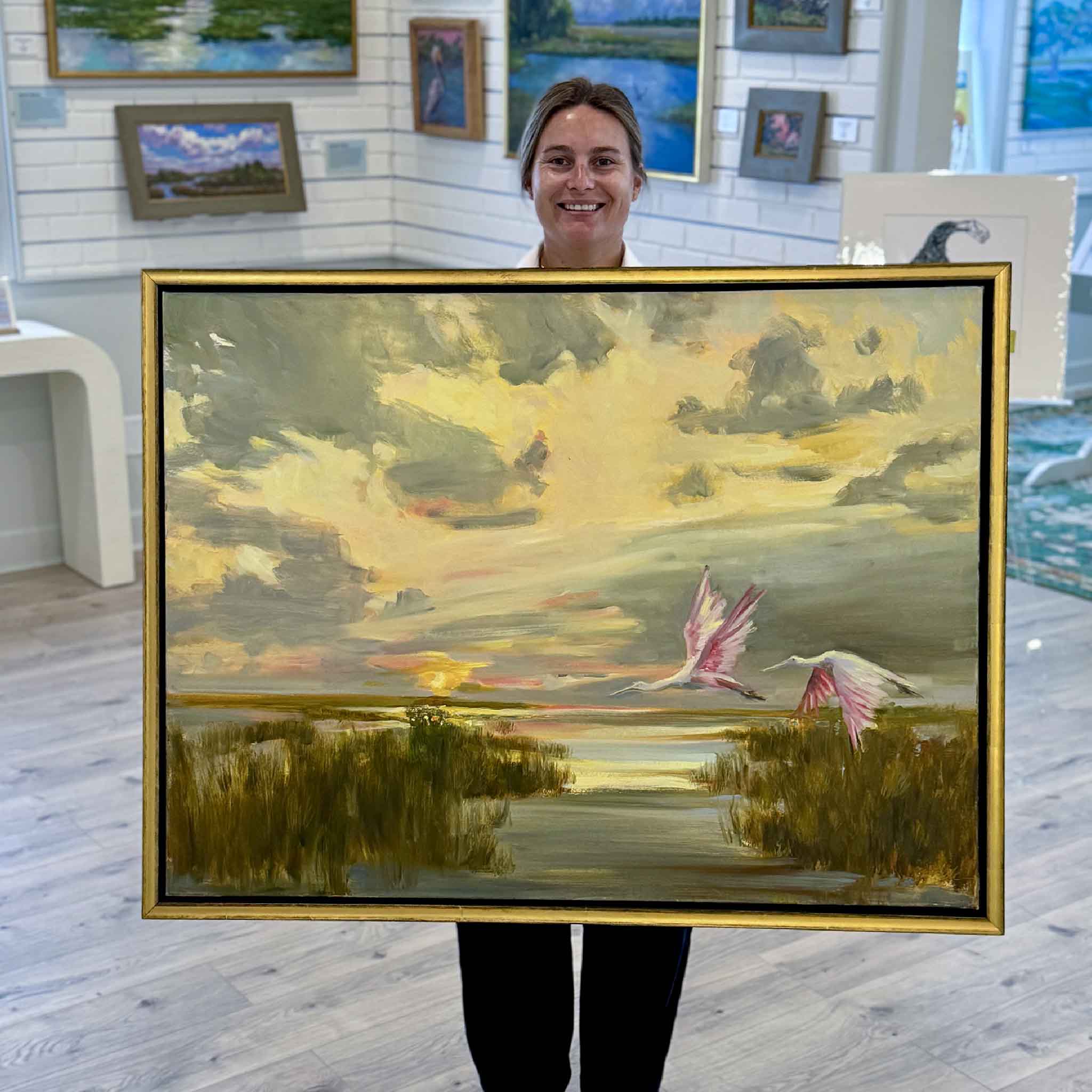 Kyra holds Cruise Control, an original framed oil painting by Helli Luck depicting roseate spoonbills gliding over a South Carolina Lowcountry marsh at sunset. Available at Gray Man Gallery in Pawleys Island, South Carolina.