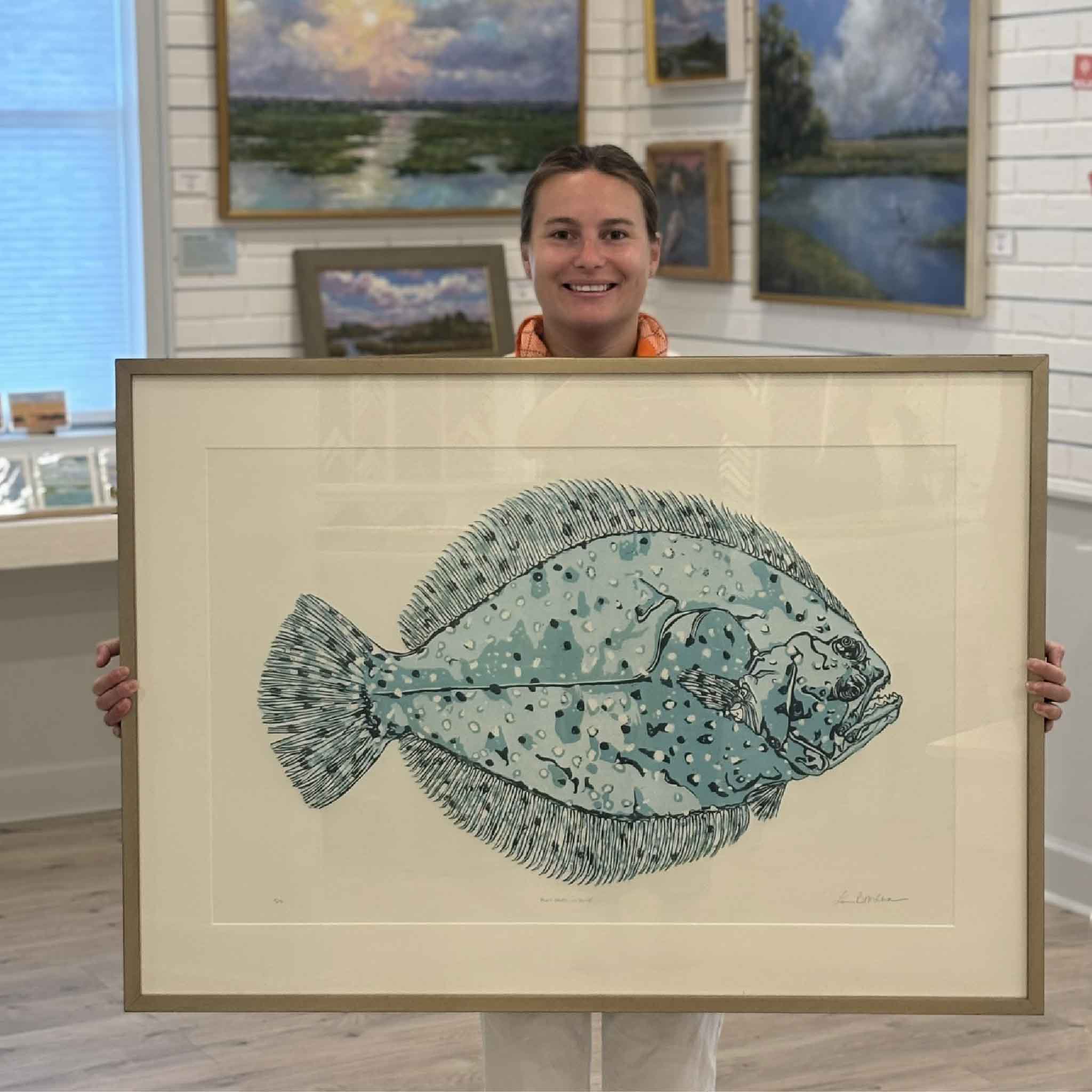 Kyra holding Flat Fred by Laurie Brownell McIntosh at Gray Man Gallery in Pawleys Island, South Carolina, showing the scale of the original framed reduction linocut featuring a blue Lowcountry flounder.