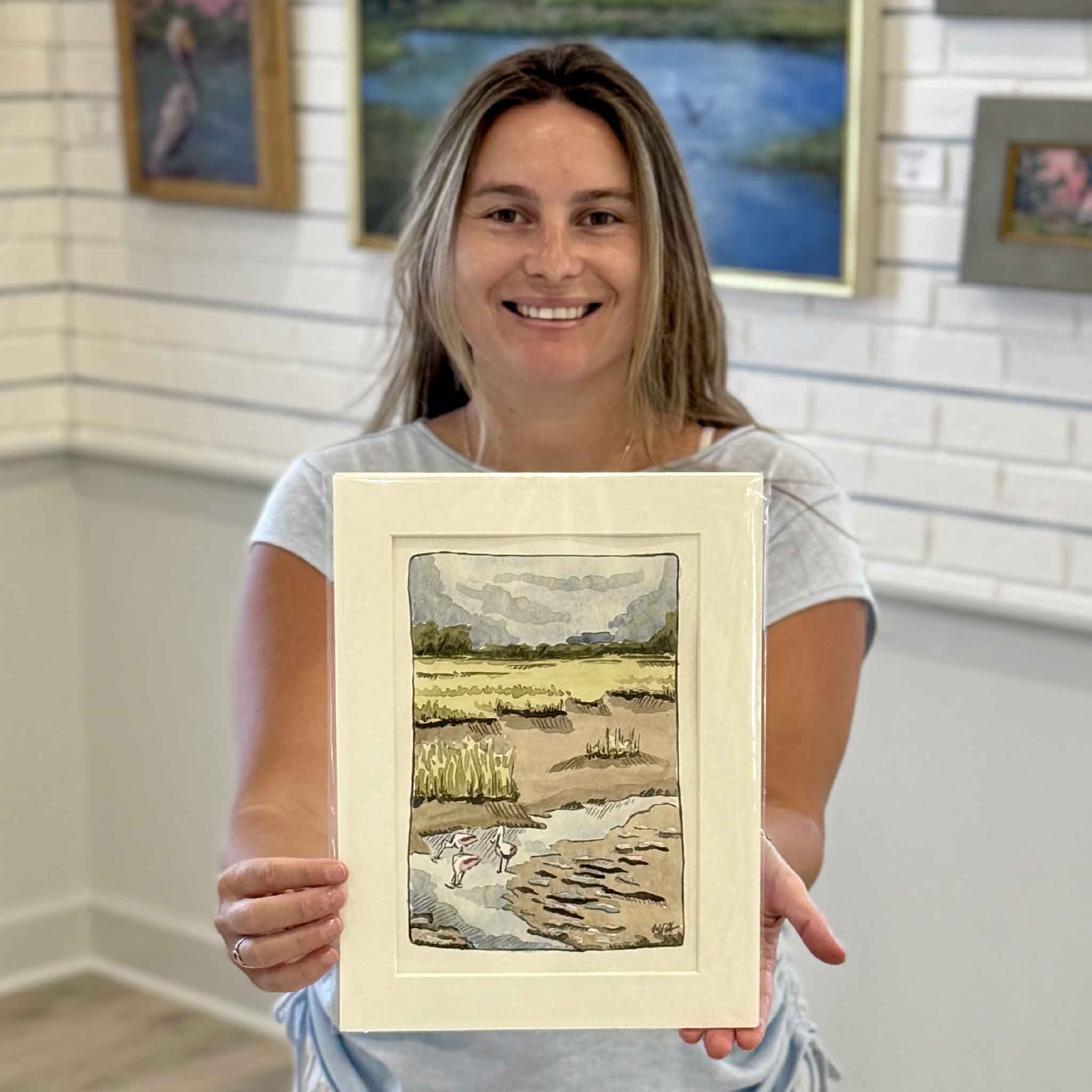 Kyra holding Following the Tide, an original watercolor by Maggie Pelton featuring roseate spoonbills feeding across a Lowcountry marsh at low tide. Professionally matted to fit a standard 9x12 frame and available at Gray Man Gallery in Pawleys Island, South Carolina.