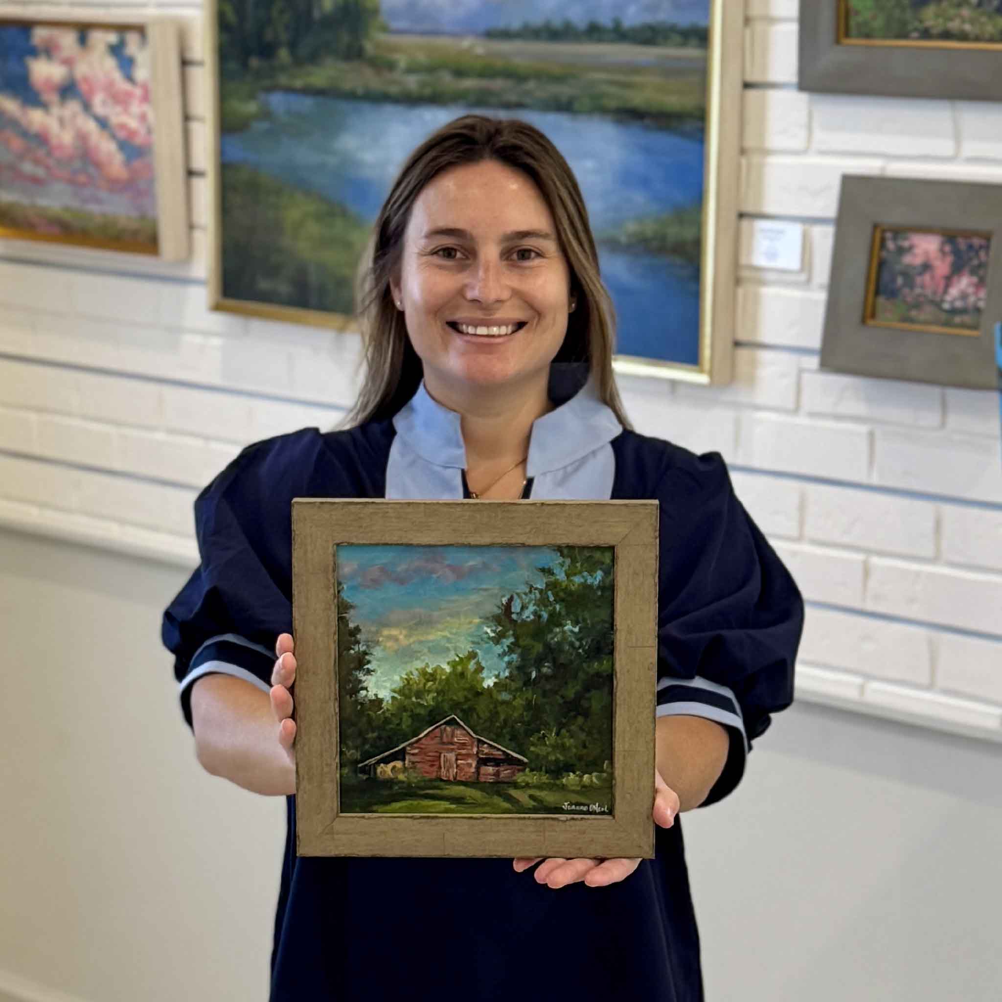 Kyra holding Grandaddy's Barn, an original oil painting by Jeanne O'Neal depicting a rustic red barn nestled among Lowcountry trees and greenery. Photographed inside Gray Man Gallery in Pawleys Island, South Carolina.