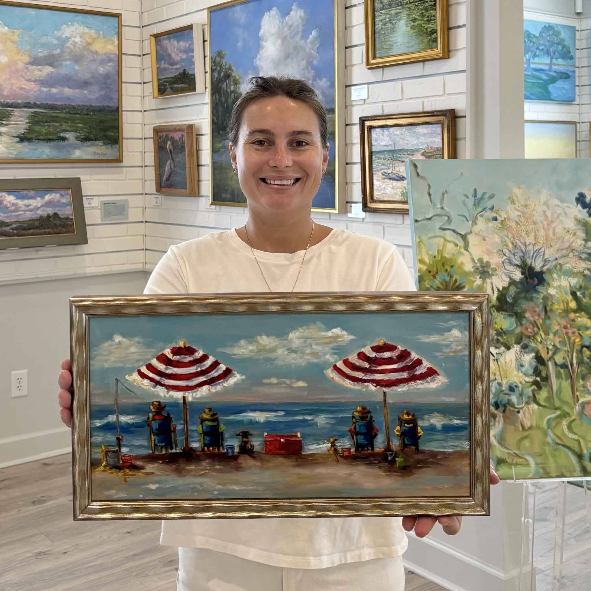 Kyra holds It's My Beach Too, an original oil painting by Jenny Reynolds, inside Gray Man Gallery in Pawleys Island, South Carolina. Framed in the artist's signature champagne frame, this vibrant coastal painting captures a relaxing day at the beach with striped umbrellas, beach chairs, dogs, fishing gear, and sparkling ocean waves, making it a joyful piece of Lowcountry wall art for beach lovers and coastal home décor.