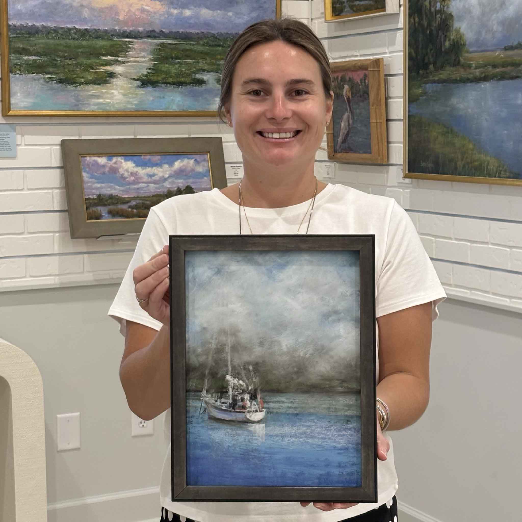 Kyra holds A Foggy Start, an original sailboat pastel by Joanne Willoughby depicting a quiet vessel anchored on calm blue water beneath a misty coastal sky. Professionally framed with Water White glass and available at Gray Man Gallery in Pawleys Island, South Carolina.