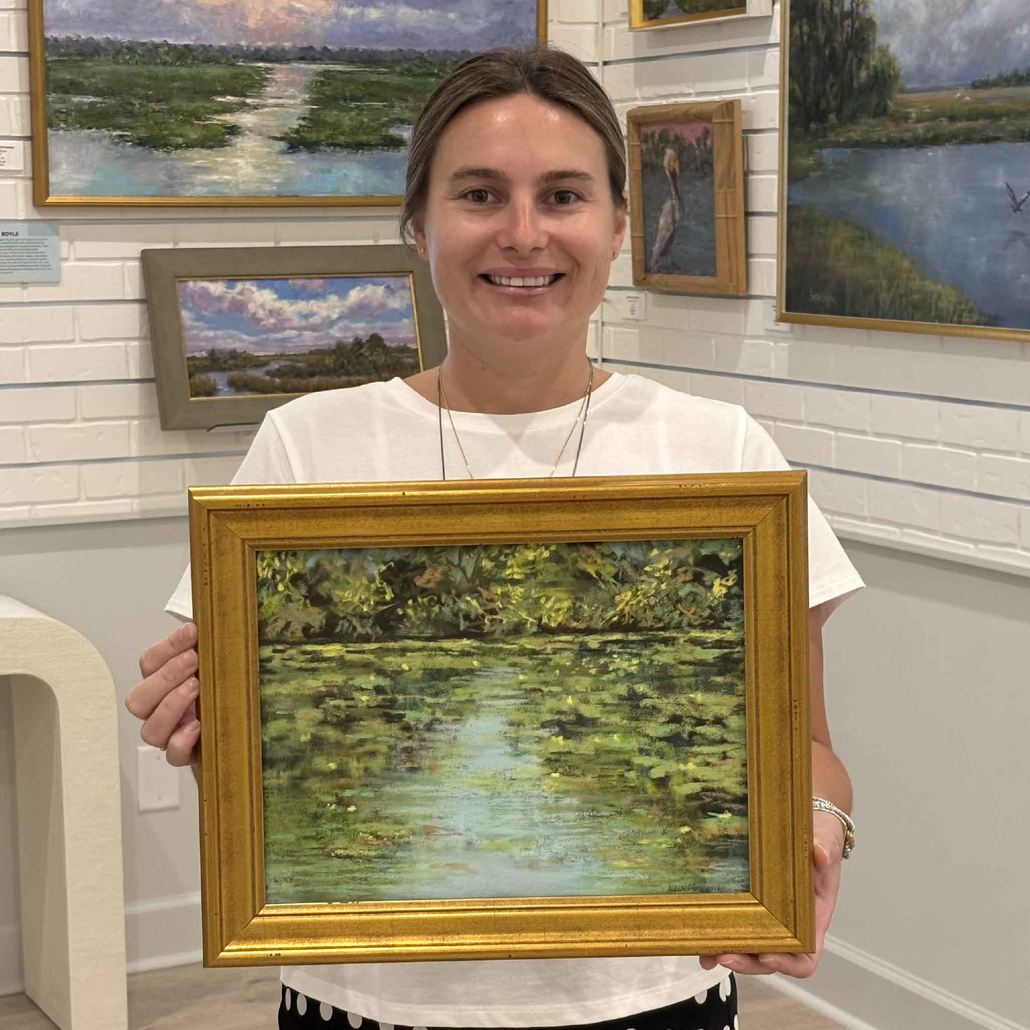 Kyra holds Afternoon at the Pond, an original soft pastel by Joanne Willoughby featuring tranquil pond reflections and lush green foliage, at Gray Man Gallery in Pawleys Island, South Carolina.