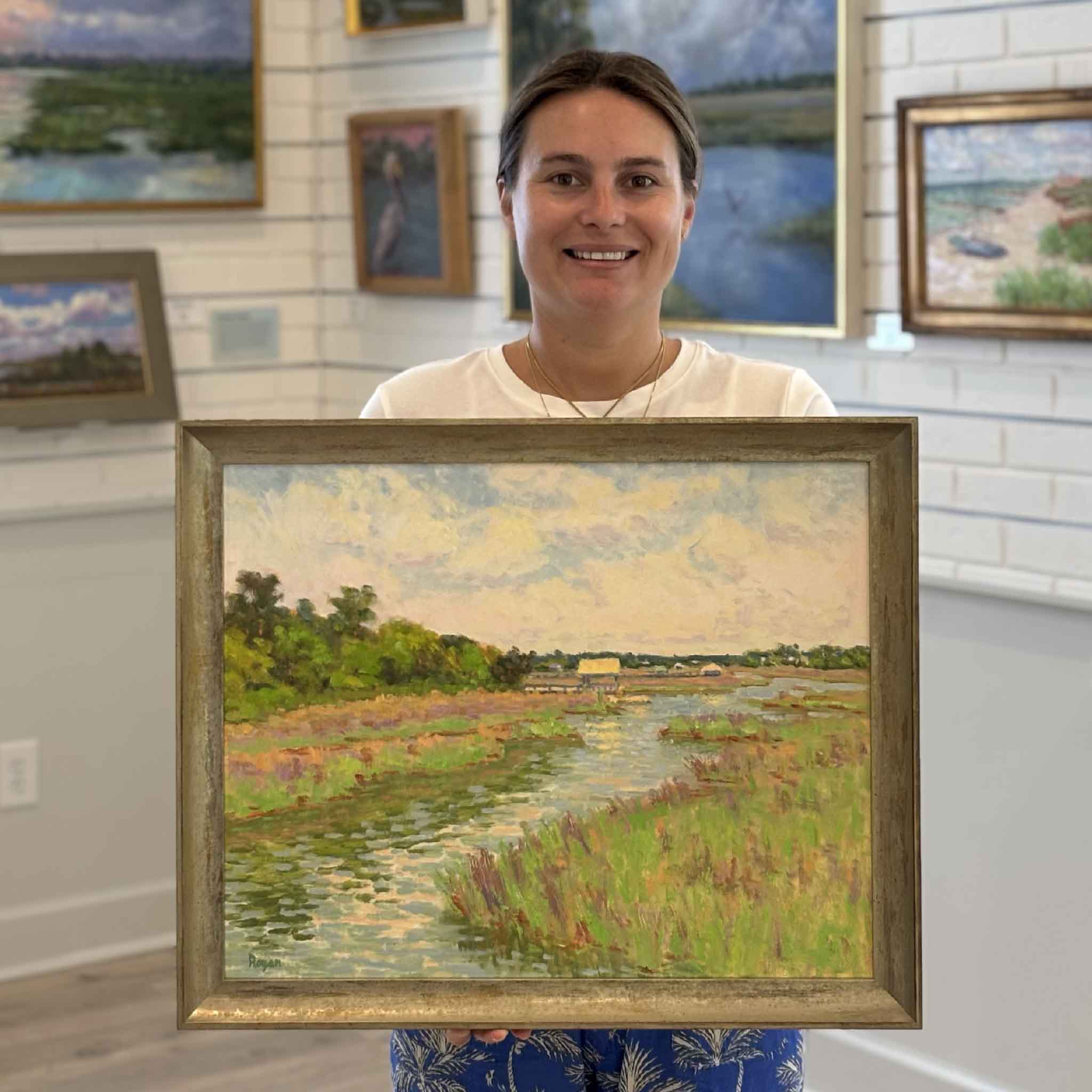 Kyra displays Spring at Litchfield, an original oil painting by Michael Rogan, at Gray Man Gallery in Pawleys Island, South Carolina. The framed Lowcountry marsh landscape captures a winding tidal creek surrounded by vibrant spring grasses beneath a luminous coastal sky, helping illustrate the artwork's scale and handcrafted weathered wood frame.
