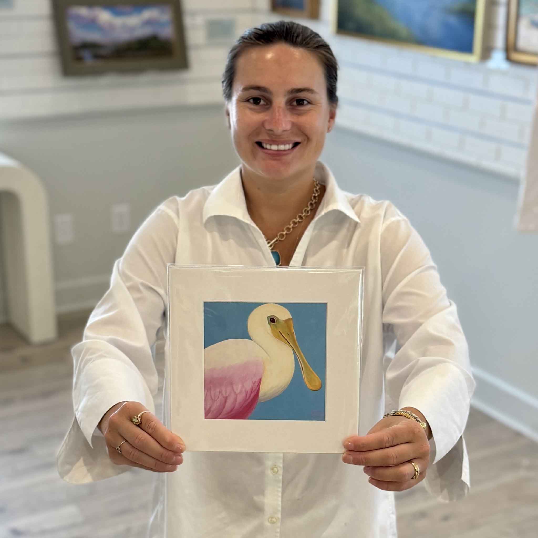 Kyra holds Roseate Spoonbill, a colorful bird print by Georgetown, South Carolina local artist Nancy Jones Lowe. Printed on 100% cotton archival fine art paper and matted to fit a standard 8×8 frame, it's available at Gray Man Gallery in Pawleys Island.
