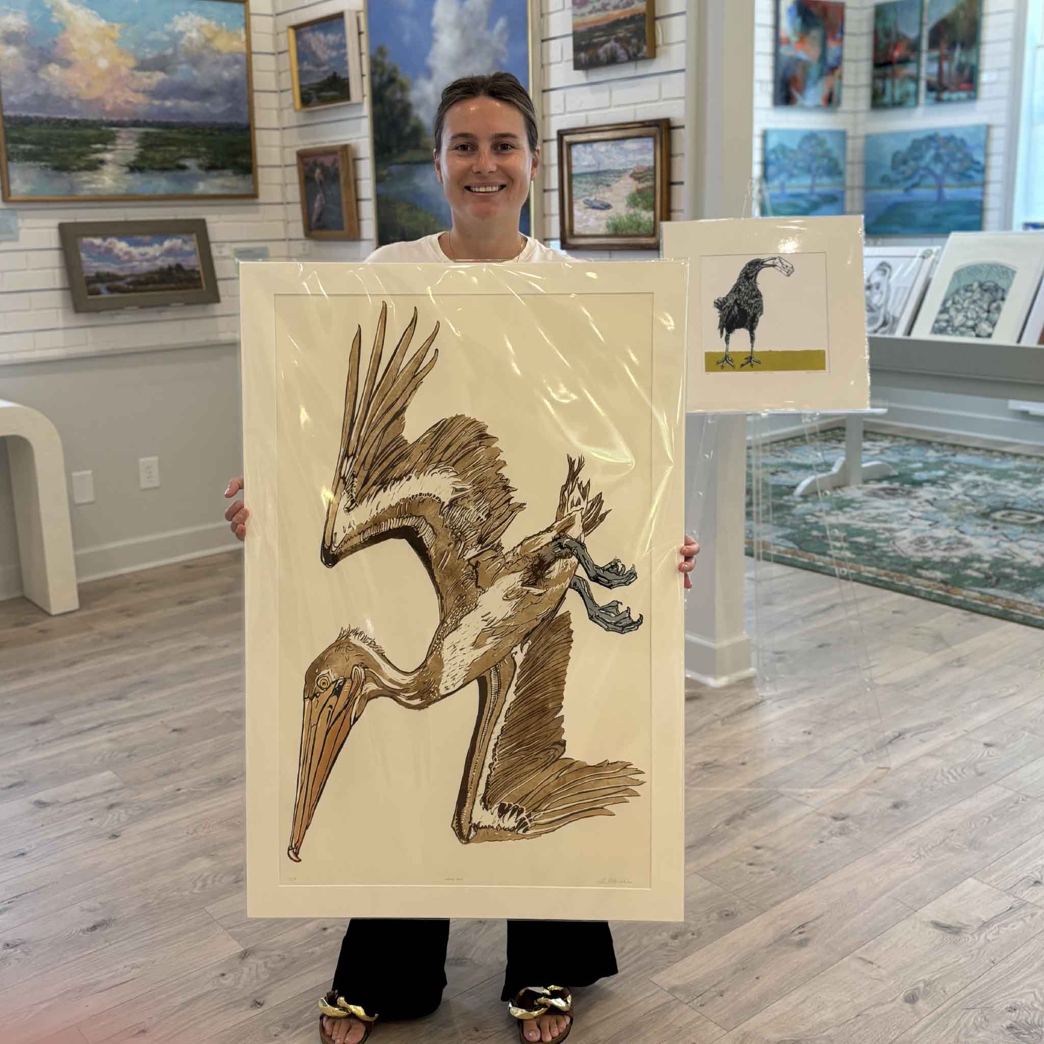 Kyra holds Nellie Peli, an original signed oil reduction linocut by Laurie Brownell McIntosh at Gray Man Gallery in Pawleys Island. Professionally matted to a standard 24x36 size, this hand-pulled original depicts a dramatic brown pelican in flight and highlights Laurie's celebrated reduction linocut process.