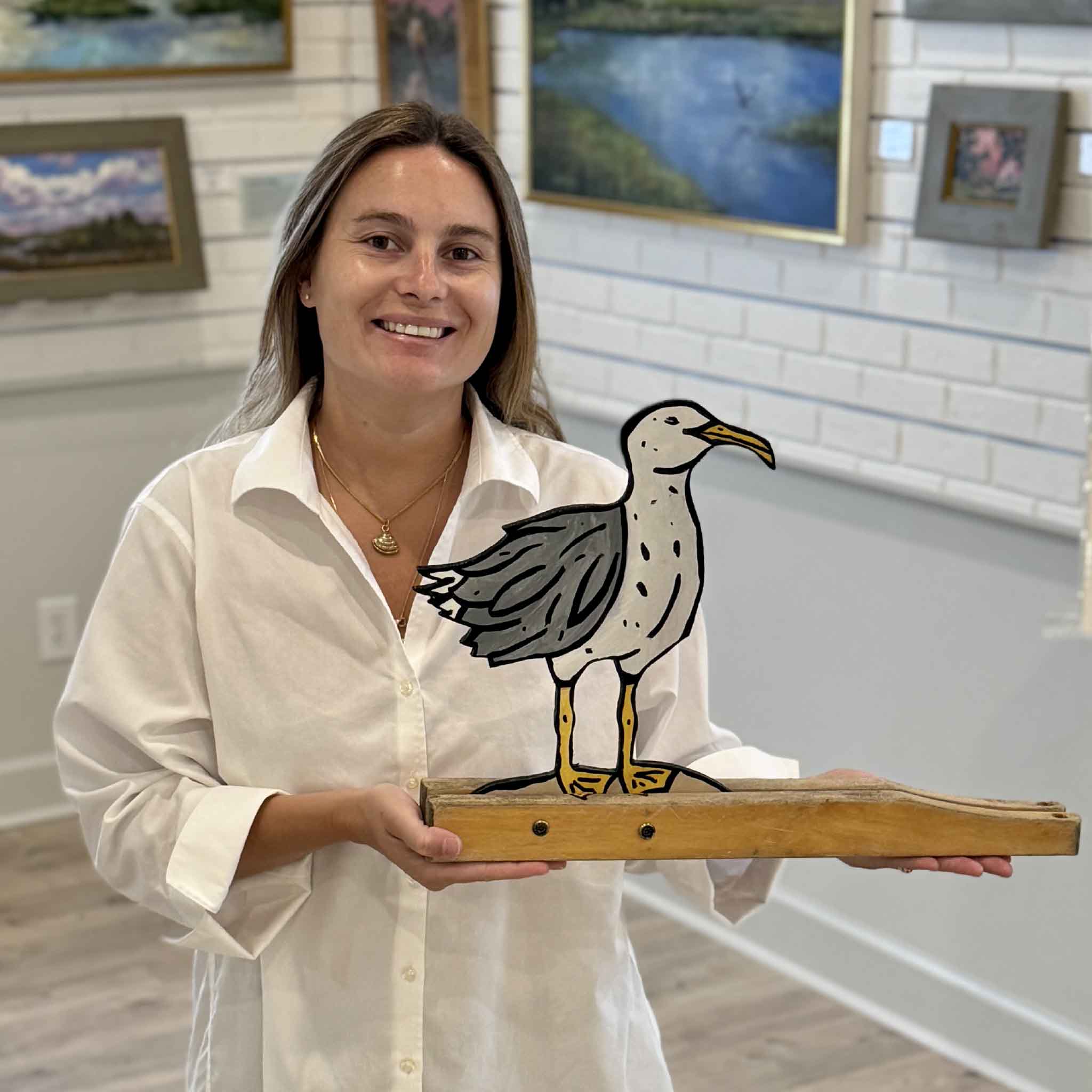 Kyra holds Seagull #9 by Nick Kawczynski inside Gray Man Gallery in Pawleys Island, South Carolina. This original handcrafted sculpture features a painted seagull cut from reclaimed marine-grade plywood and mounted to a salvaged wooden base, highlighting the scale and character of this one-of-a-kind coastal artwork.