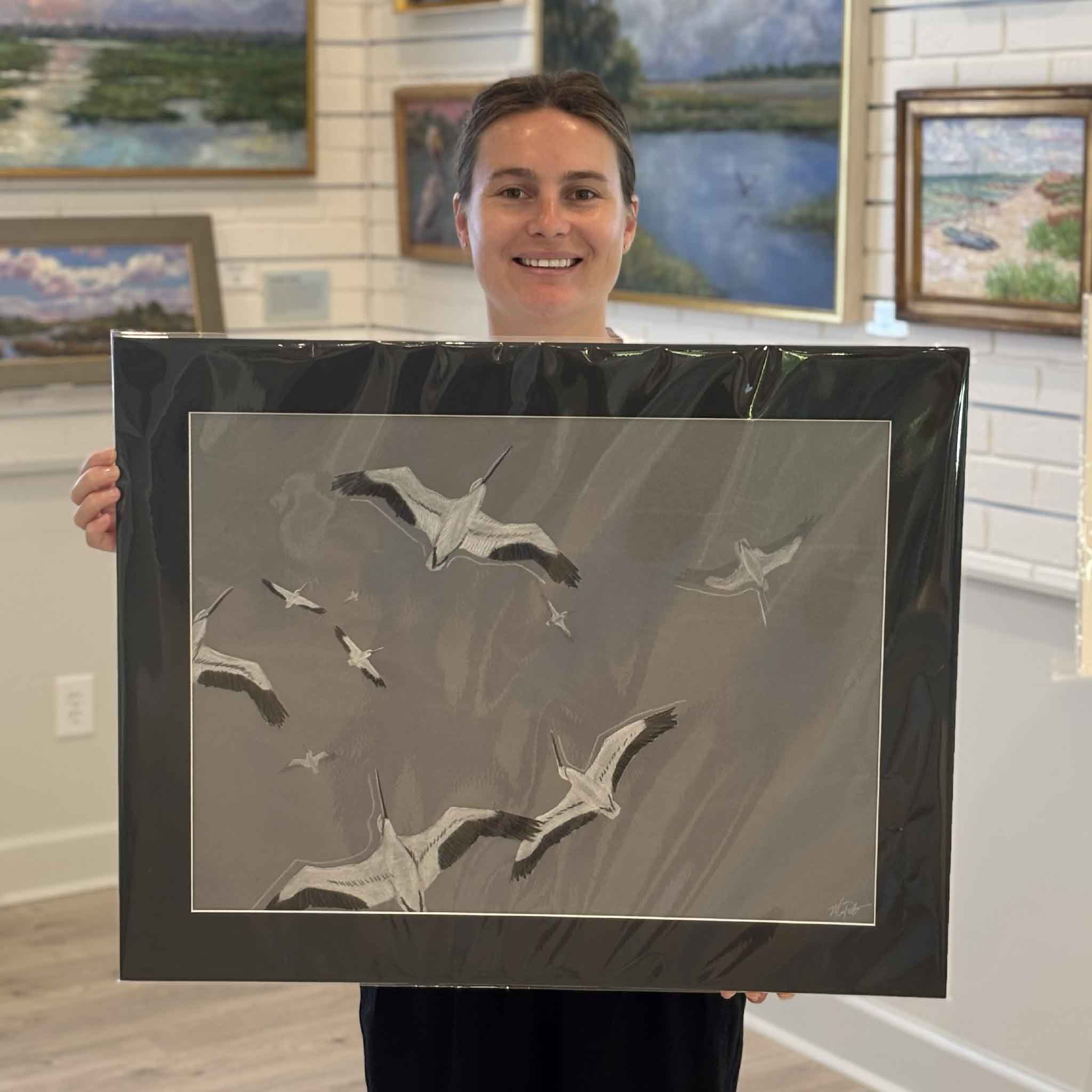 Kyra holds Passing Through, an original mixed-media artwork by Maggie Pelton at Gray Man Gallery in Pawleys Island. This one-of-a-kind original features layered tracing paper, graphite, and ink to create remarkable depth as a flock of wood storks moves across the Lowcountry sky. Professionally matted to fit a standard 22x28 frame.