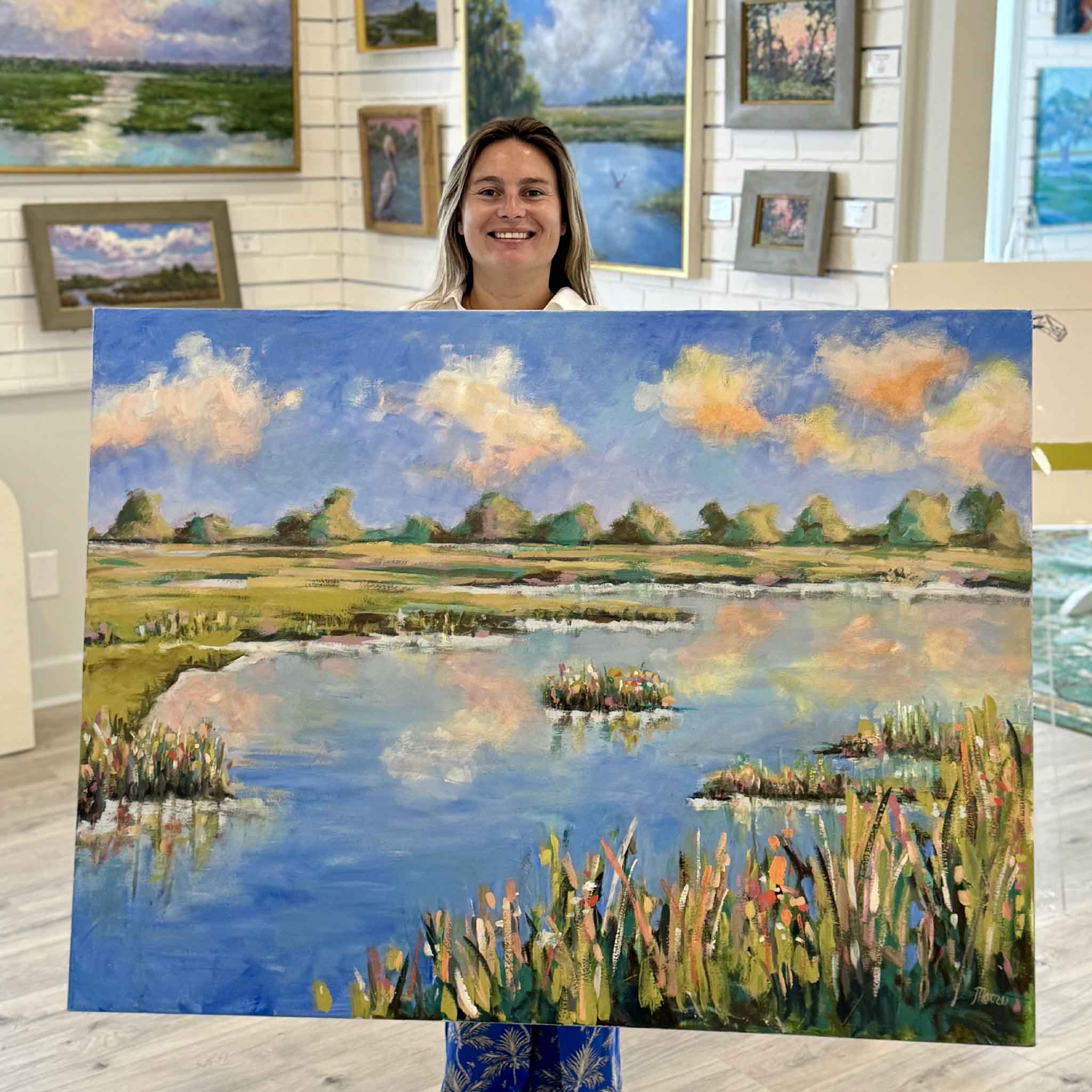 Kyra holding Shabby Chic by local artist Susan Moore at Gray Man Gallery in Pawleys Island, SC. This 48x36 original acrylic painting captures the beauty of the Lowcountry marsh near North Inlet with colorful marsh grasses, reflective water, and a bright Carolina sky filled with soft pink clouds.