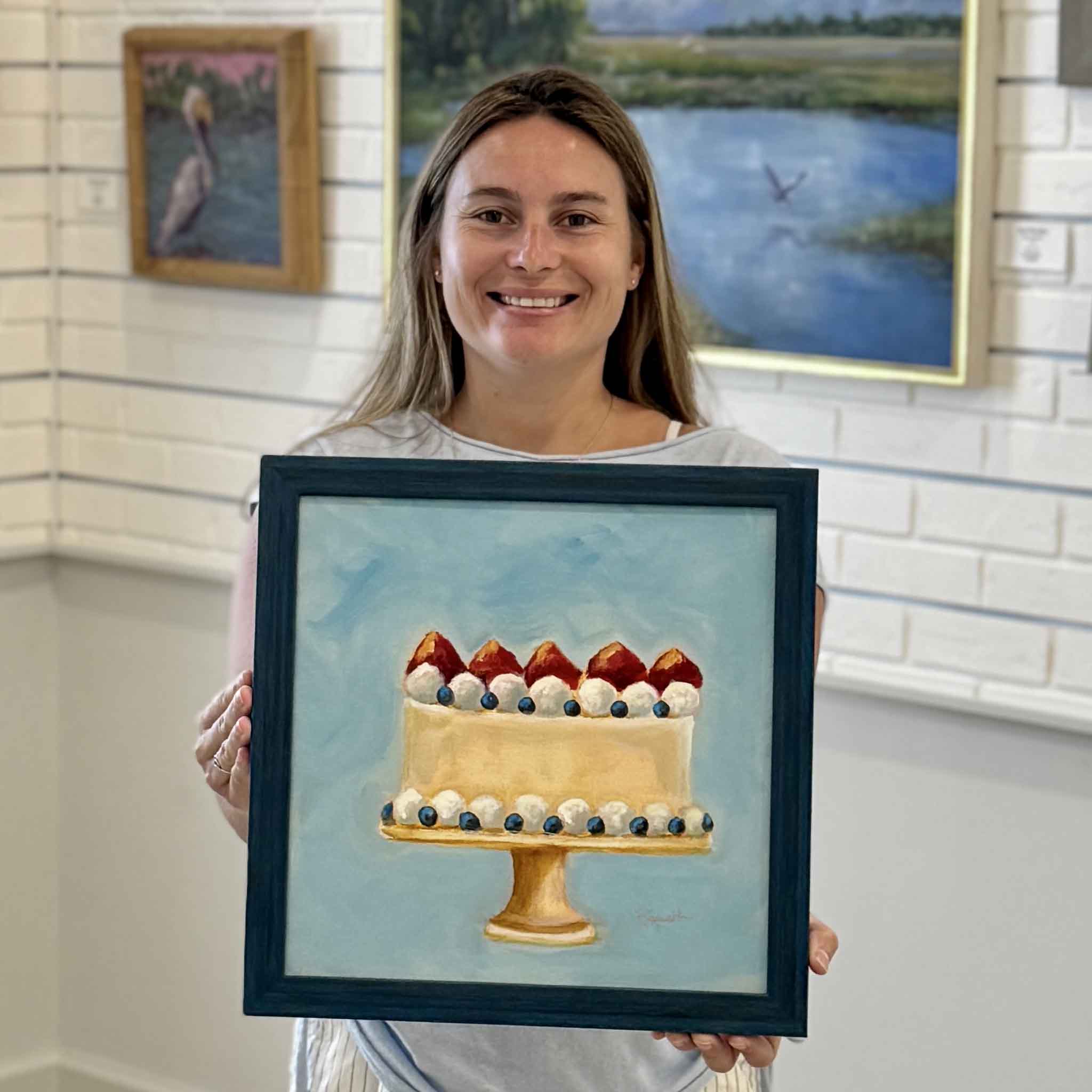 Kyra holding Tastes Like Summer Pound Cake by local artist Jenny Reynolds at Gray Man Gallery in Pawleys Island, South Carolina. This original oil painting from the Southern Sweets collection features a strawberry pound cake topped with strawberries, whipped cream, and blueberries in a bright blue frame.