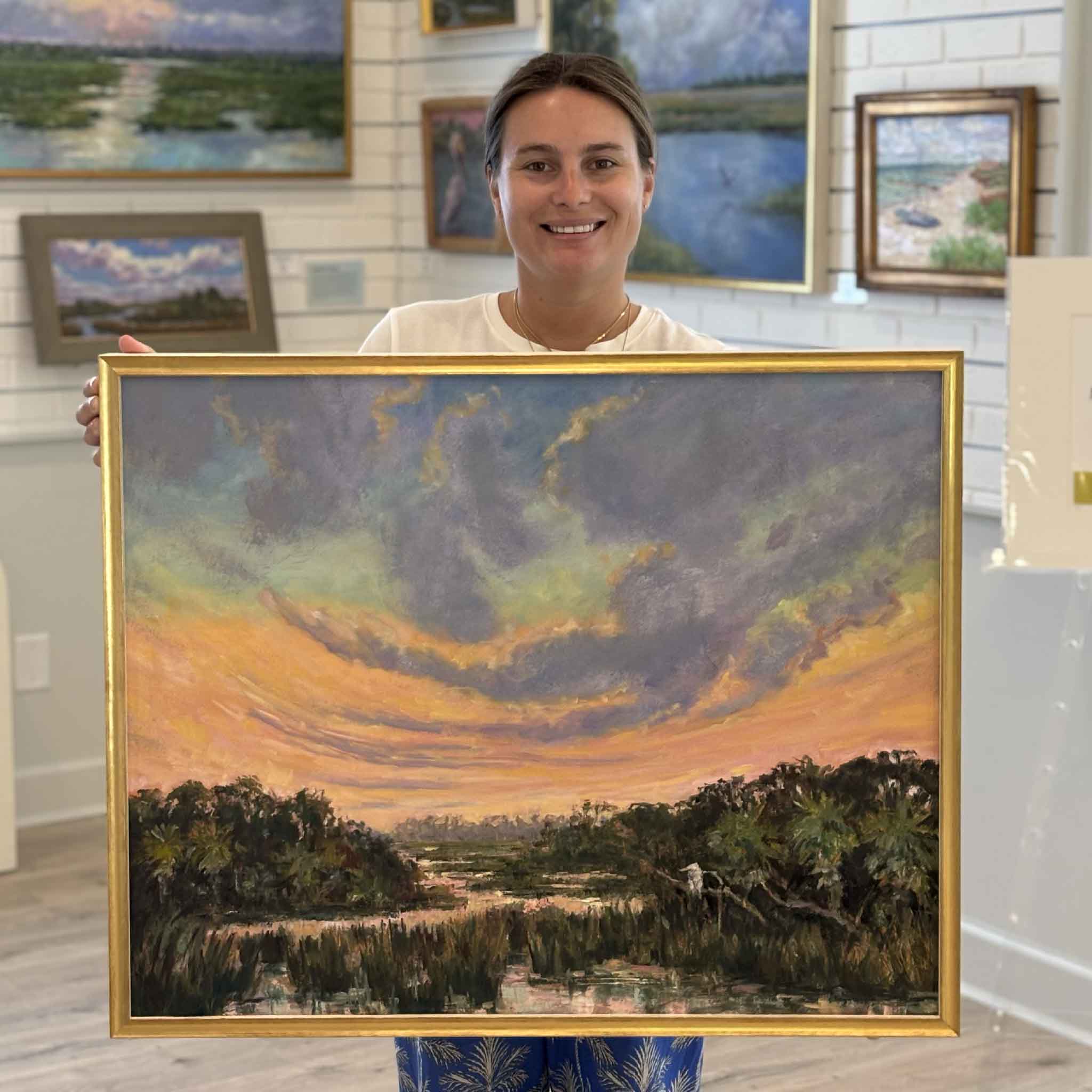 Kyra holds The Day Softens, an original oil painting by South Carolina artist Jeanne O'Neal at Gray Man Gallery in Pawleys Island. Framed in Jeanne's signature gold frame, the painting features an expansive sunset sky over the Lowcountry marsh with rich pastel clouds reflected in the winding tidal creek.