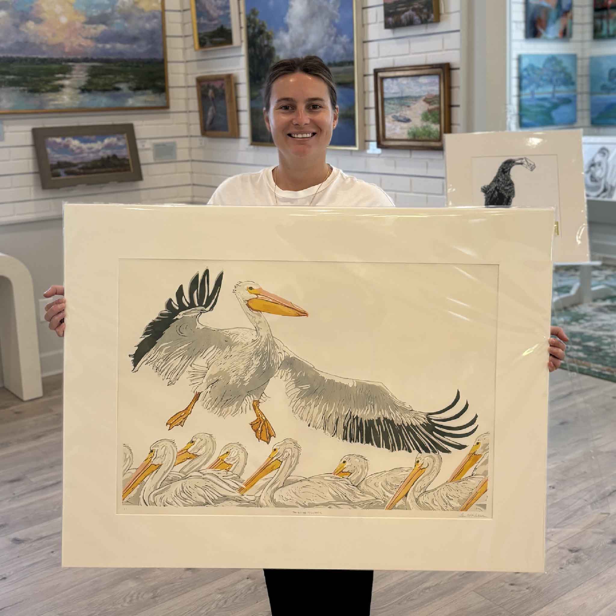 Kyra holds The White Pelicans, an original signed oil reduction linocut by Laurie Brownell McIntosh at Gray Man Gallery in Pawleys Island. Professionally matted to fit a standard 36x24 frame, this hand-pulled original depicts a flock of American white pelicans with one bird lifting into flight and demonstrates the artist's celebrated reduction linocut technique.