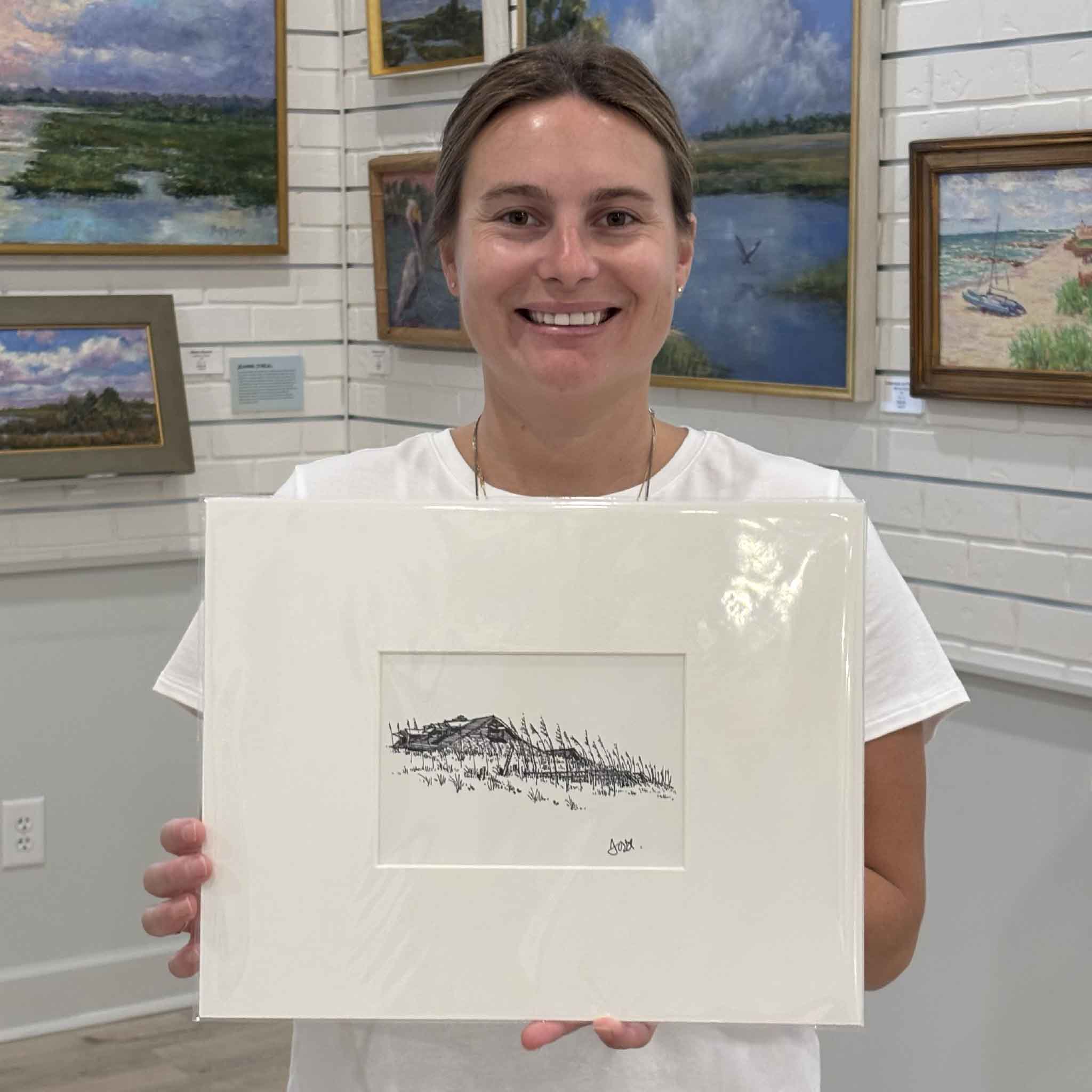 Kyra holds Todd Halamka's matted fine art print, Pawleys Beach House with Sea Oats, inside Gray Man Gallery in Pawleys Island, South Carolina. The image showcases the scale of the professionally matted coastal print, which fits a standard ready-made frame without custom framing and celebrates the timeless beauty of a Pawleys Island beach house tucked behind sea oats.
