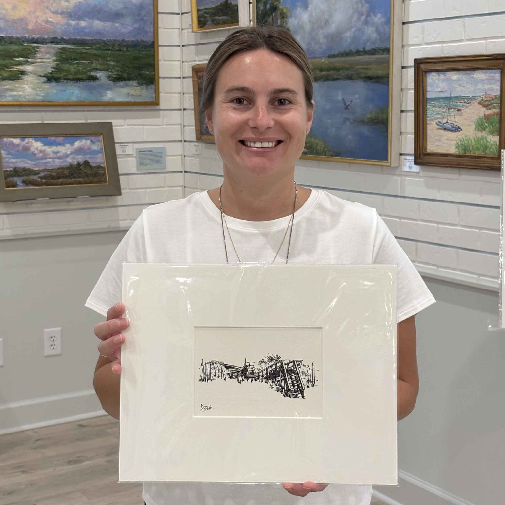 Kyra holds Todd Halamka's Pawleys Beach House Boardwalk print inside Gray Man Gallery in Pawleys Island, South Carolina. Professionally matted to a standard 11x14 size, this detailed pen-and-ink print captures the classic wooden boardwalk leading from a Pawleys Island beach house through the dunes and is ready for any standard 11x14 frame.