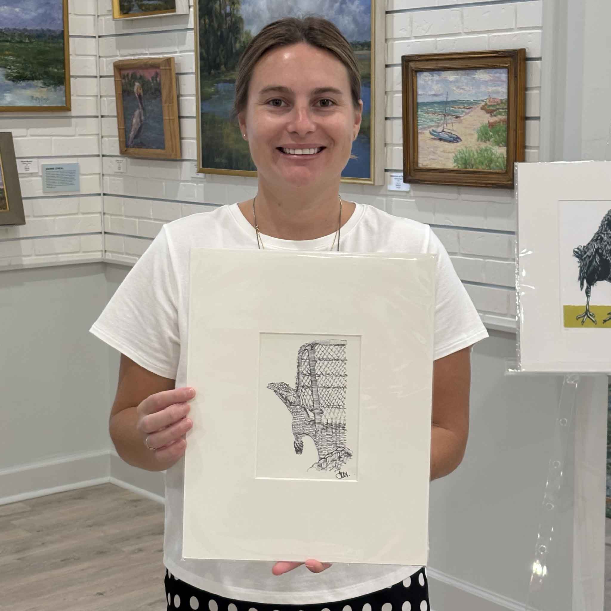 Kyra holds Pawleys Island Wicker Chair, a matted fine art print by Pawleys Island artist Todd Halamka, inside Gray Man Gallery in Pawleys Island, South Carolina. The image shows the scale of the professionally matted print featuring Todd's detailed pen-and-ink drawing of the island's iconic wicker porch chair, ready for a standard frame without custom framing.
