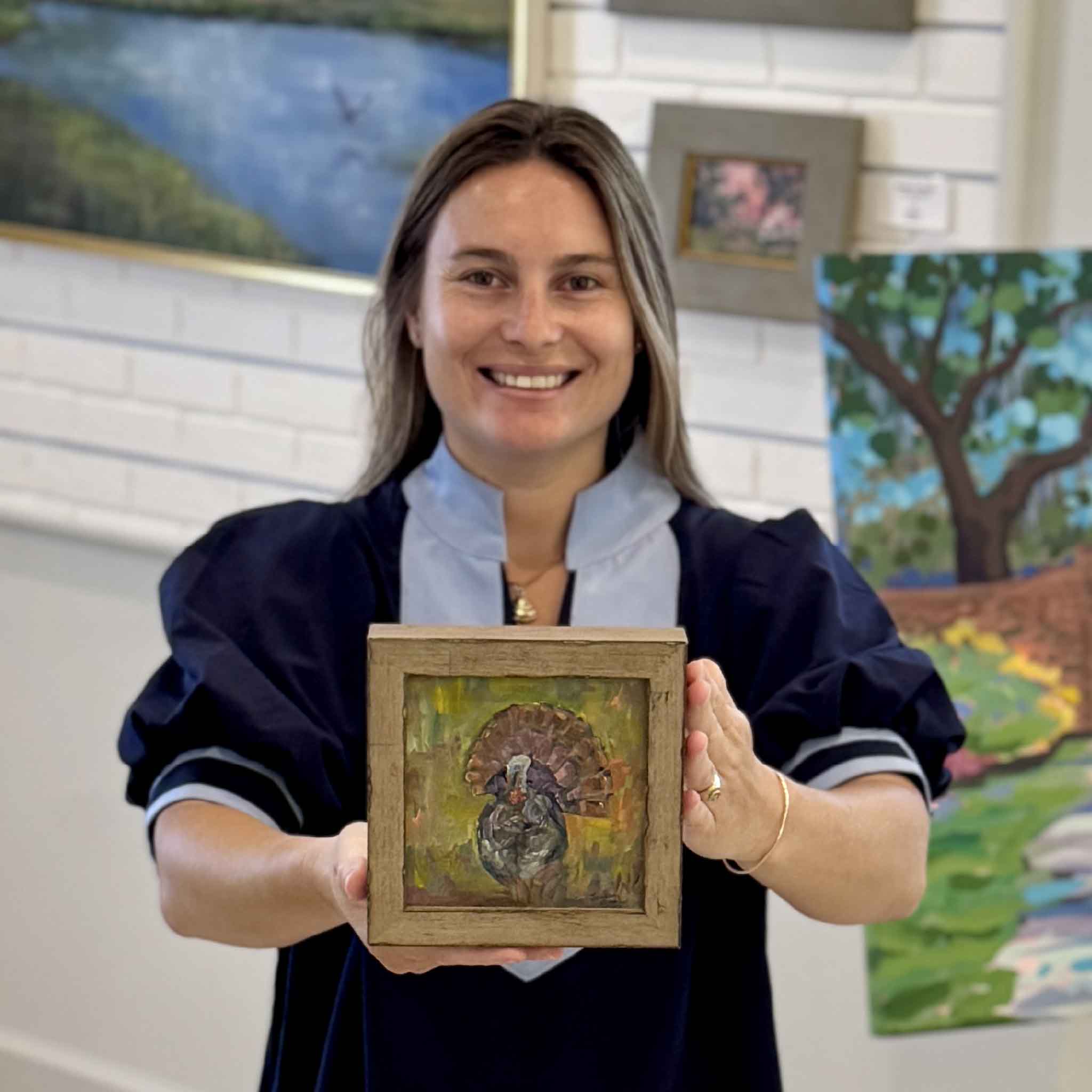 Kyra holding Turkey Season, an original oil painting by local artist Jeanne O'Neal featuring a colorful wild turkey painted in expressive brushstrokes and framed in rustic wood. Photographed inside Gray Man Gallery in Pawleys Island, South Carolina.