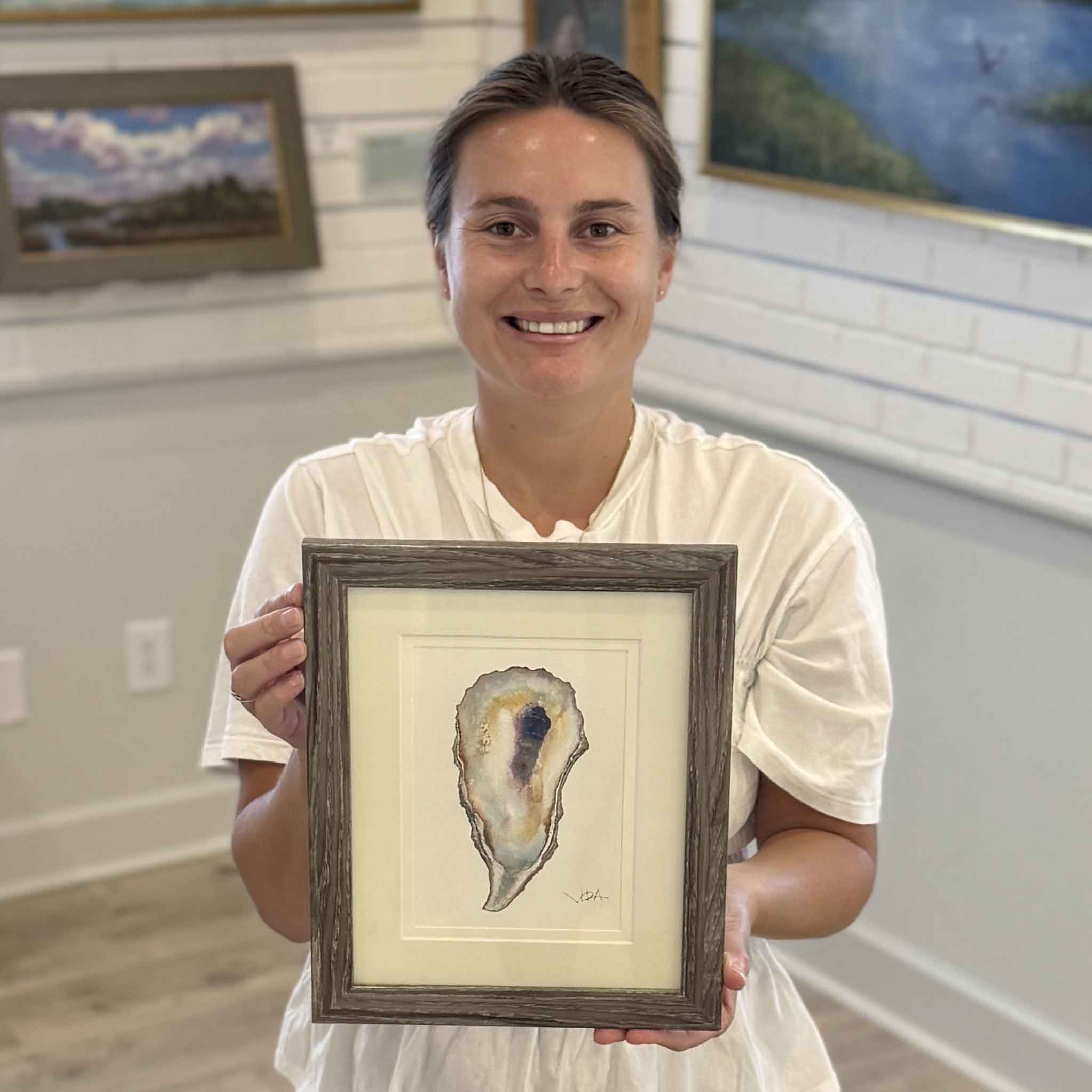 Kyra displays Pawleys Oyster Shell IX, an original watercolor by local Pawleys Island artist Vida Miller. Professionally double matted and framed, this one-of-a-kind oyster shell painting features luminous pearl whites, warm honey tones, soft coastal blues, and a dramatic indigo center, available at Gray Man Gallery in Pawleys Island, South Carolina.