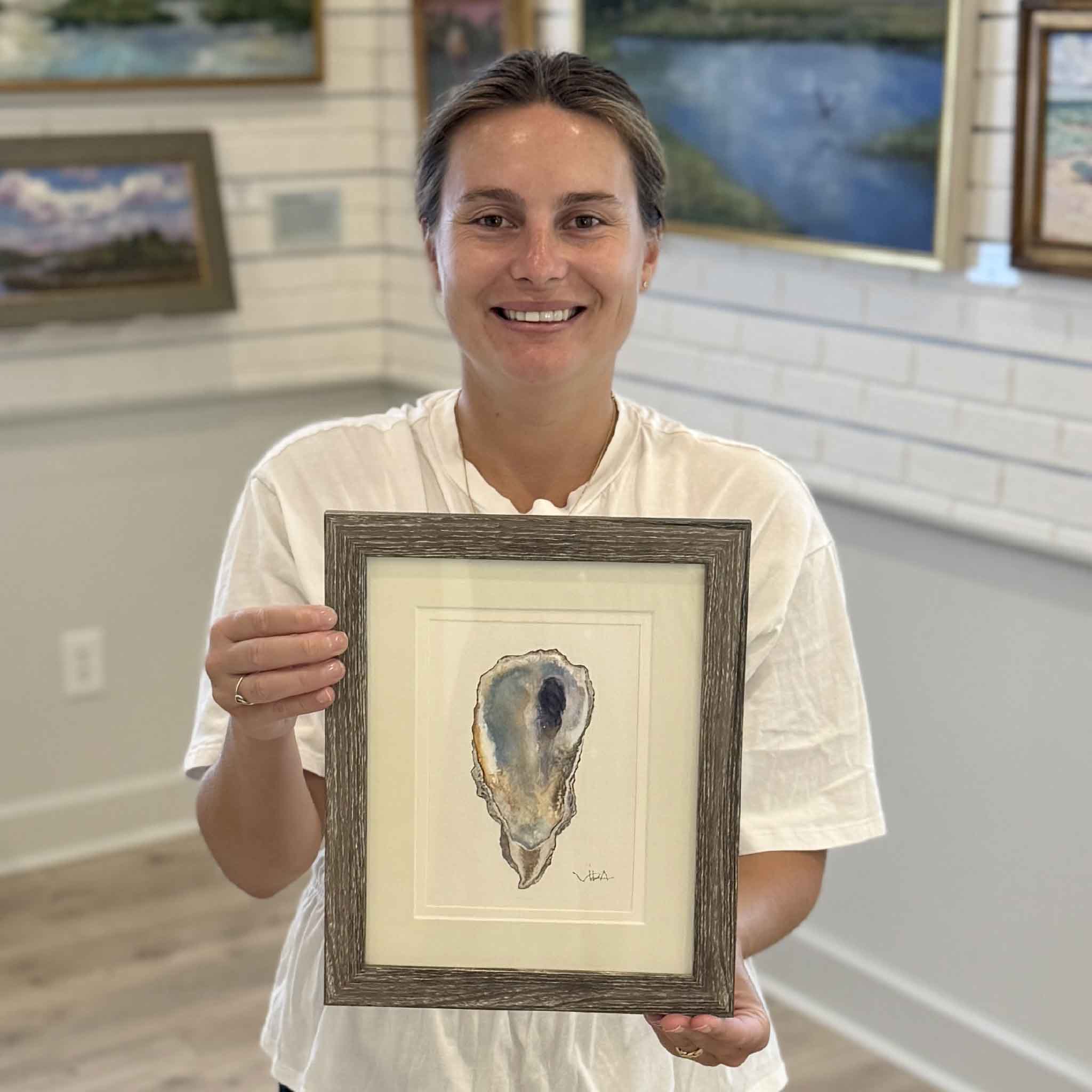 Kyra displays Pawleys Oyster Shell VII, an original watercolor by local artist Vida Miller. Beautifully double matted and framed, this one-of-a-kind coastal painting features soft blue-gray tones, creamy ivory highlights, and a distinctive violet interior inspired by an oyster shell collected along Pawleys Island, available at Gray Man Gallery.
