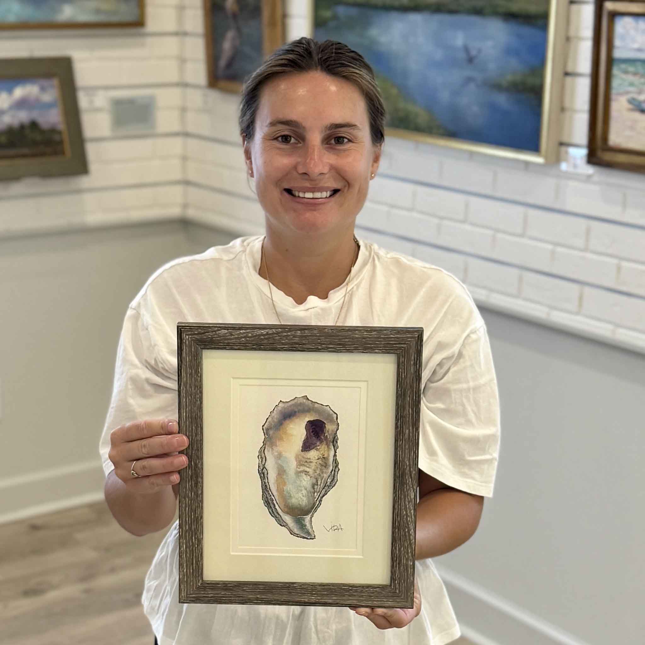 Kyra holds Pawleys Oyster Shell VIII, an original watercolor by Vida Miller featuring soft pearl whites, pale aqua blues, warm golden tones, and delicate lavender accents. This professionally double matted and framed original coastal painting is available at Gray Man Gallery in Pawleys Island, South Carolina.