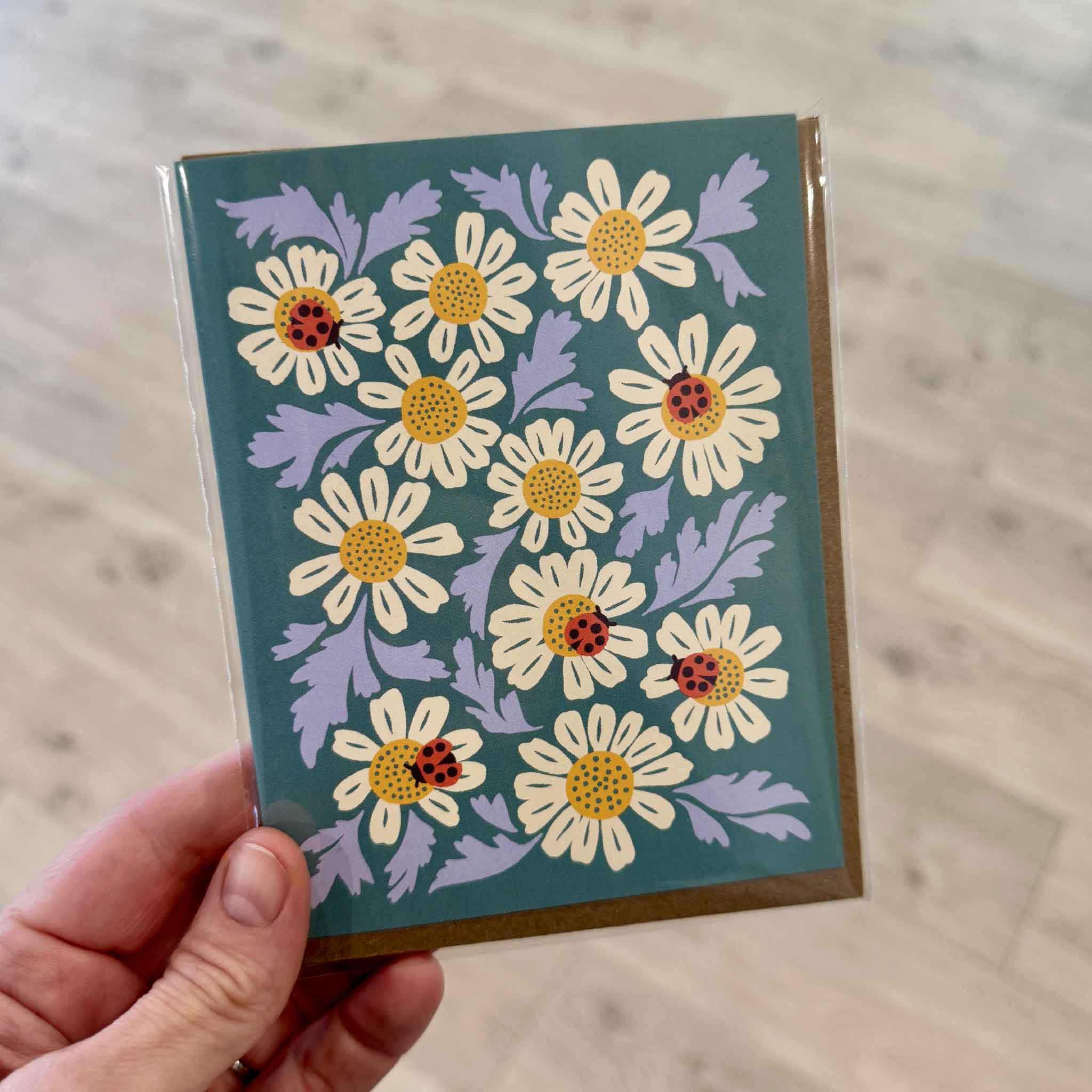 Customer holding the Ladybugs & Chamomile Greeting Card by Nuthatch Studio inside Gray Man Gallery in Pawleys Island, South Carolina. This charming botanical greeting card features cheerful white chamomile flowers accented with bright red ladybugs on a soft teal background, printed on premium 100% recycled paper and paired with a recycled kraft envelope for an eco-friendly gift or thoughtful handwritten note.