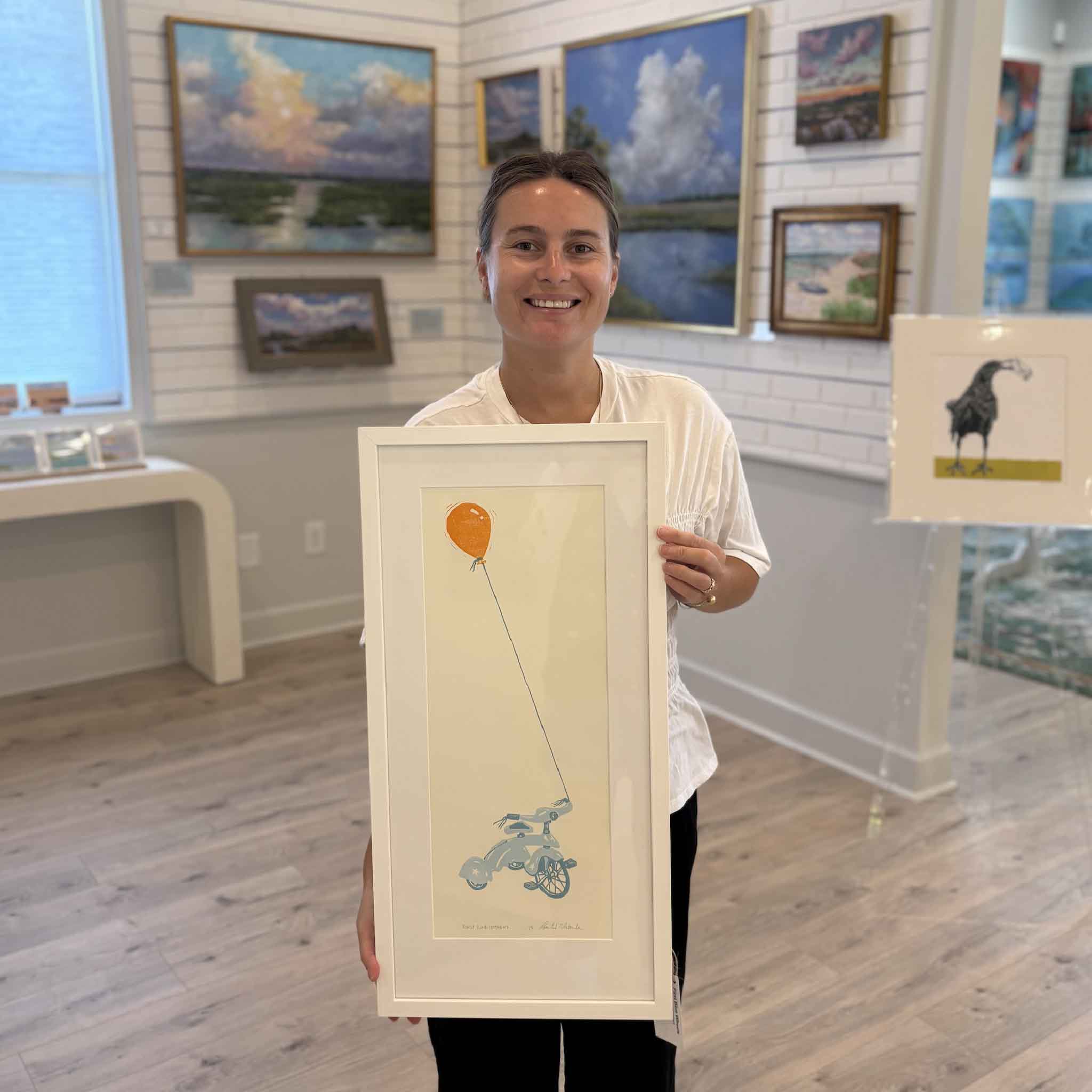 Kyra holds First Blue Wheels, an original hand-pulled reduction linocut by South Carolina artist Laurie McIntosh, at Gray Man Gallery in Pawleys Island. This whimsical original print features a blue tricycle tethered to a bright orange balloon, showcasing the artwork's scale, custom white frame, and playful color palette. Available exclusively at Gray Man Gallery, this one-of-a-kind linocut celebrates childhood wonder through Laurie McIntosh's signature reduction printmaking process.