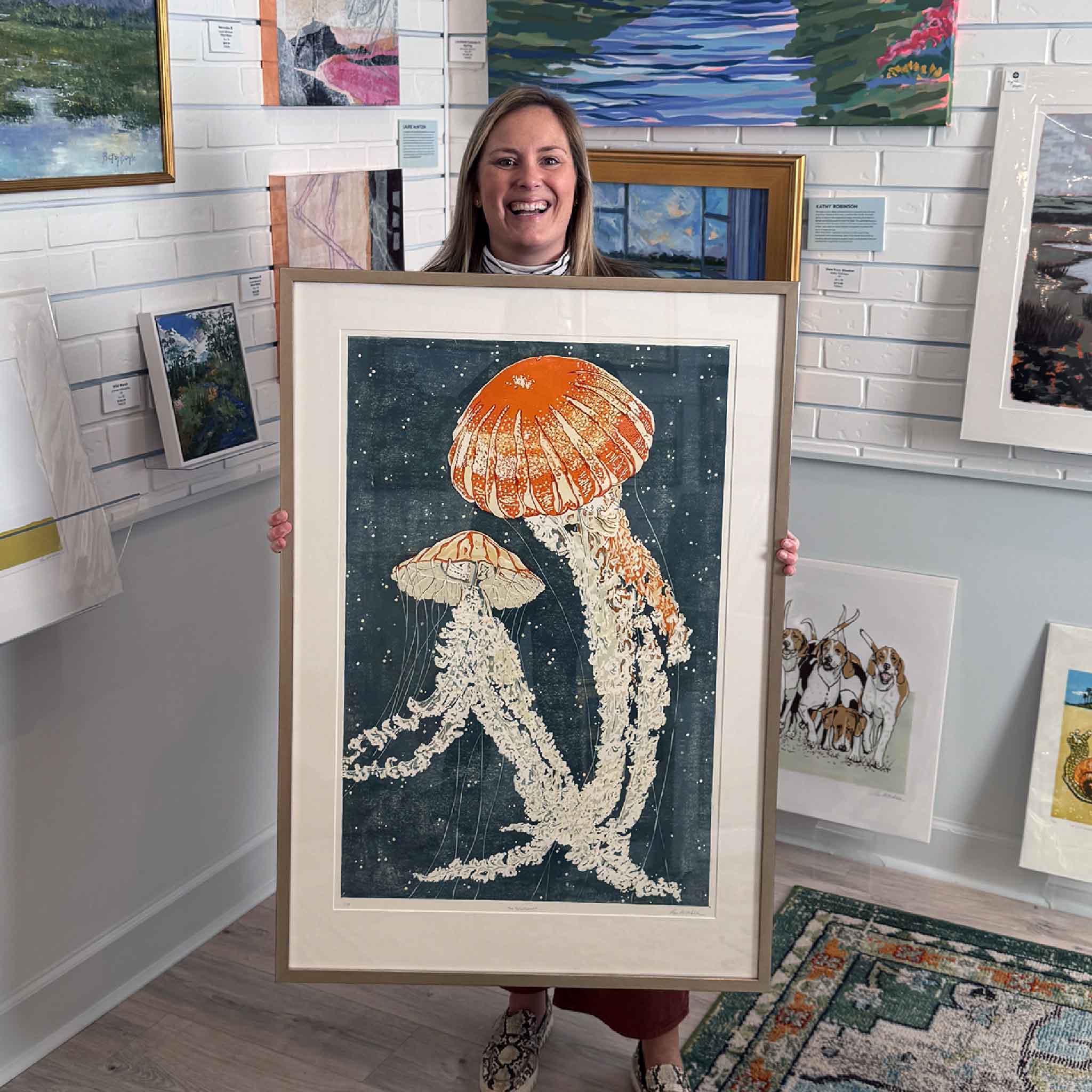 Smiling Bordy holding local artist Laurie McIntosh's framed “Jelly Dance” reduction linocut print at Gray Man Gallery in Pawleys Island, South Carolina, featuring vibrant orange jellyfish on deep blue background, coastal fine art and printmaking