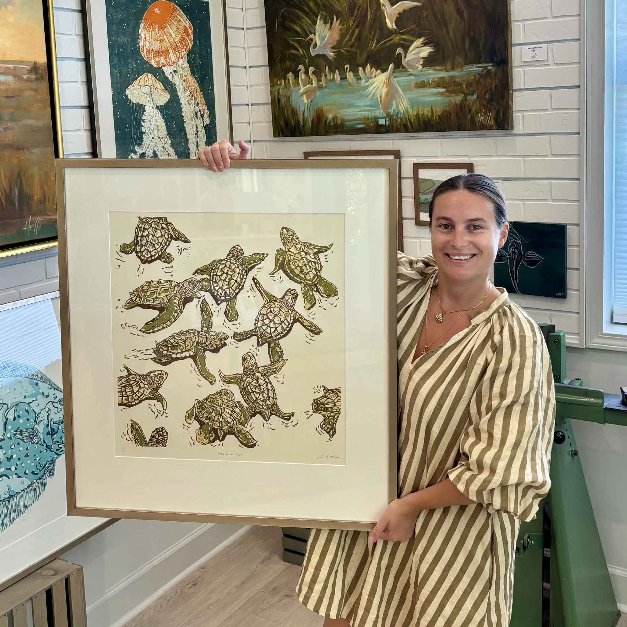 Kyra holding Let’s Follow Her by Laurie McIntosh, an original hand-pulled reduction linocut featuring a lively group of young sea turtles, beautifully matted and framed in the artist’s signature champagne frame inside Gray Man Gallery in Pawleys Island, South Carolina.