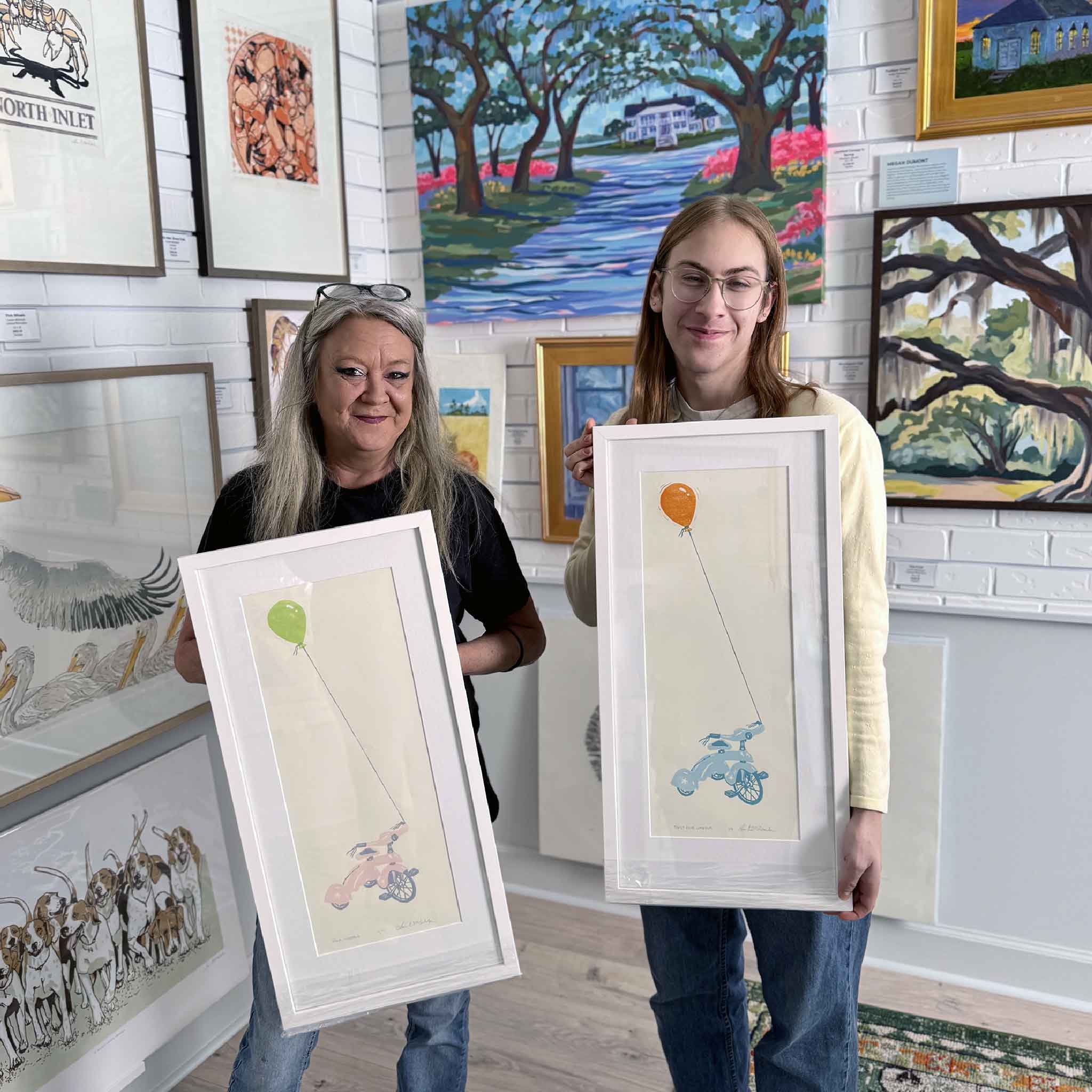 Charlotte and Alex holding Laurie McIntosh's Pink Wheels and First Blue Wheels linocut prints inside Gray Man Gallery in Pawleys Island.