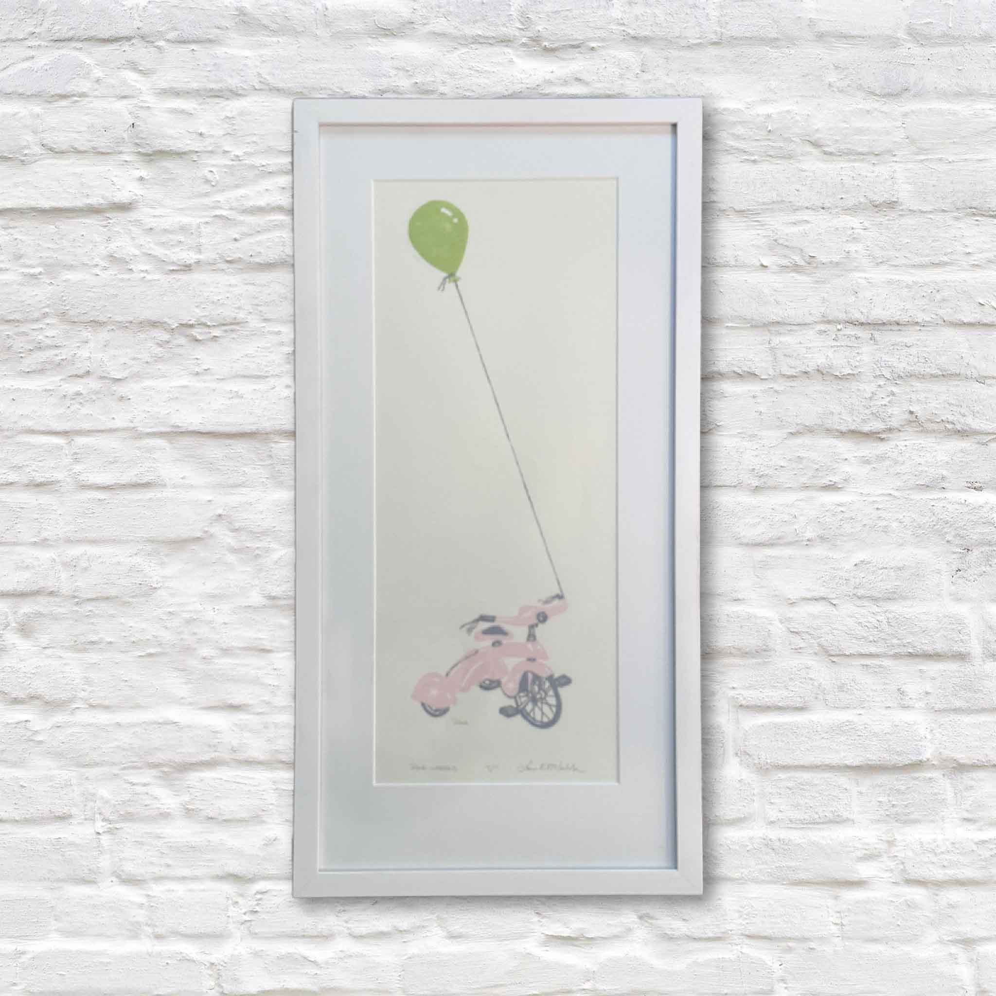 Original Pink Wheels linocut by local artist Laurie McIntosh featuring a soft pink tricycle tethered to a bright green balloon, professionally framed and displayed at Gray Man Gallery in Pawleys Island, South Carolina.